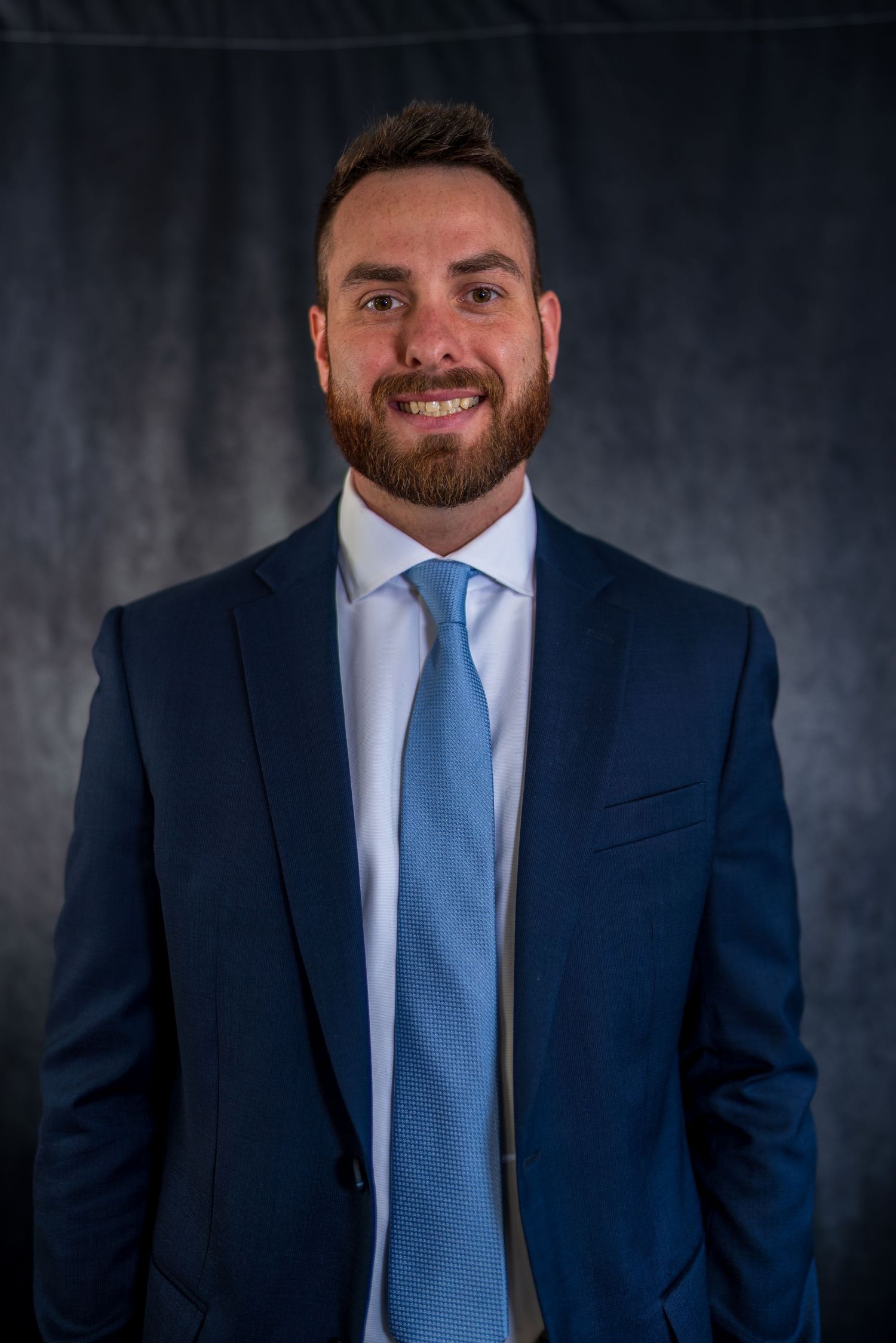 Corporate portrait series showing man in navy suit and blue tie against dark backdrop.