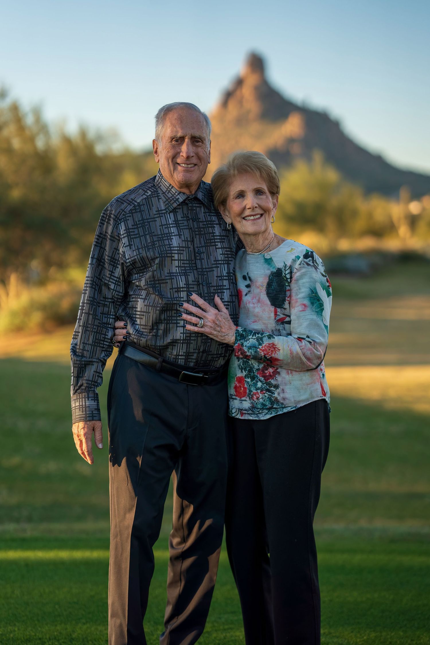 Elderly couple embracing on grassy field with mountain backdrop at sunset.