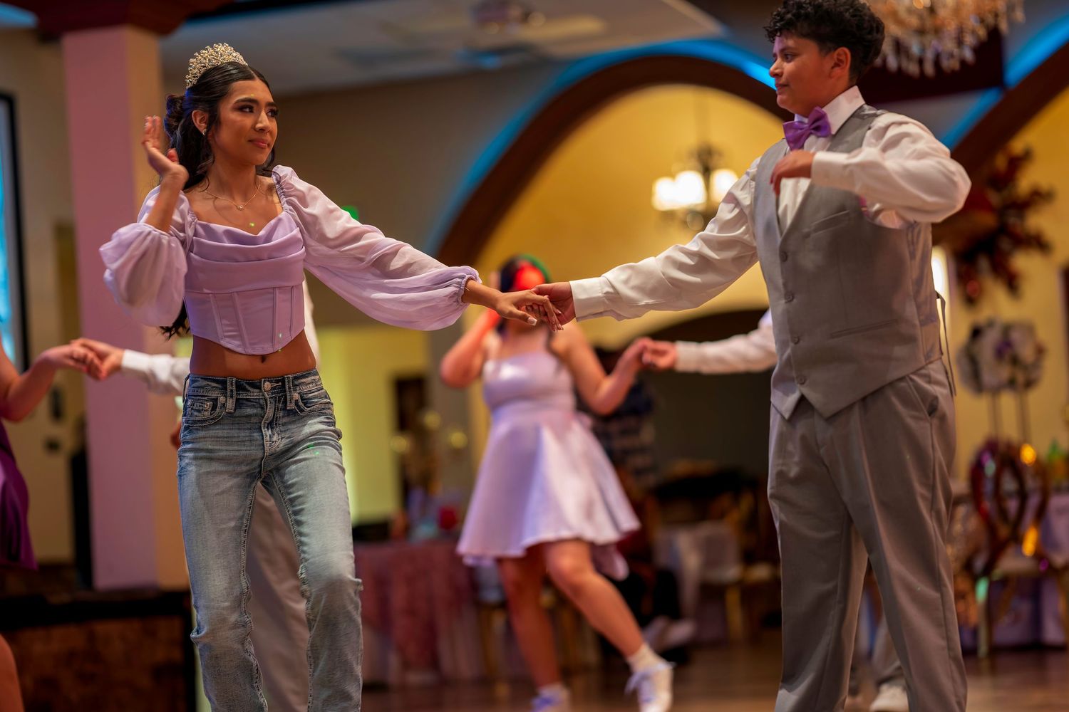 Two teens dance together at a formal event, girl in lavender top and jeans, boy in gray suit with purple bow tie.