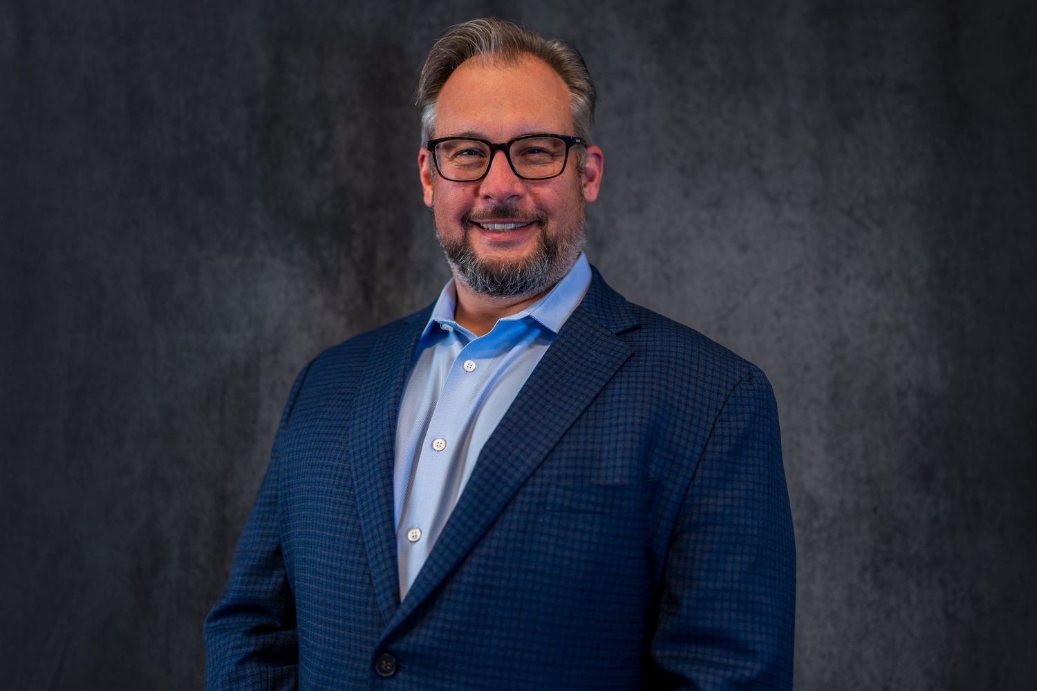 Professional headshot featuring blue blazer against dark studio background.