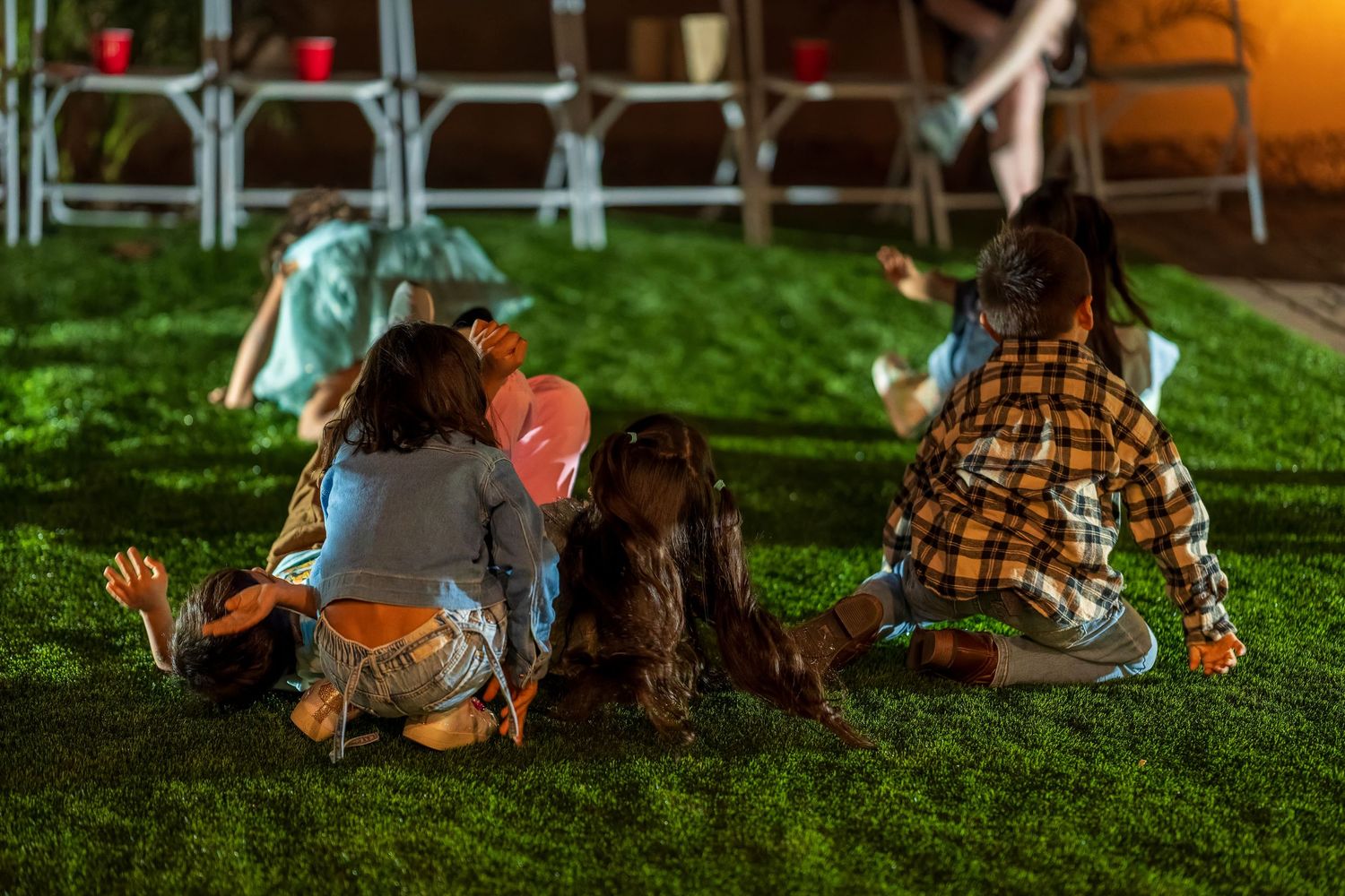 Children and adults sitting on grass near folding chairs at an outdoor event.