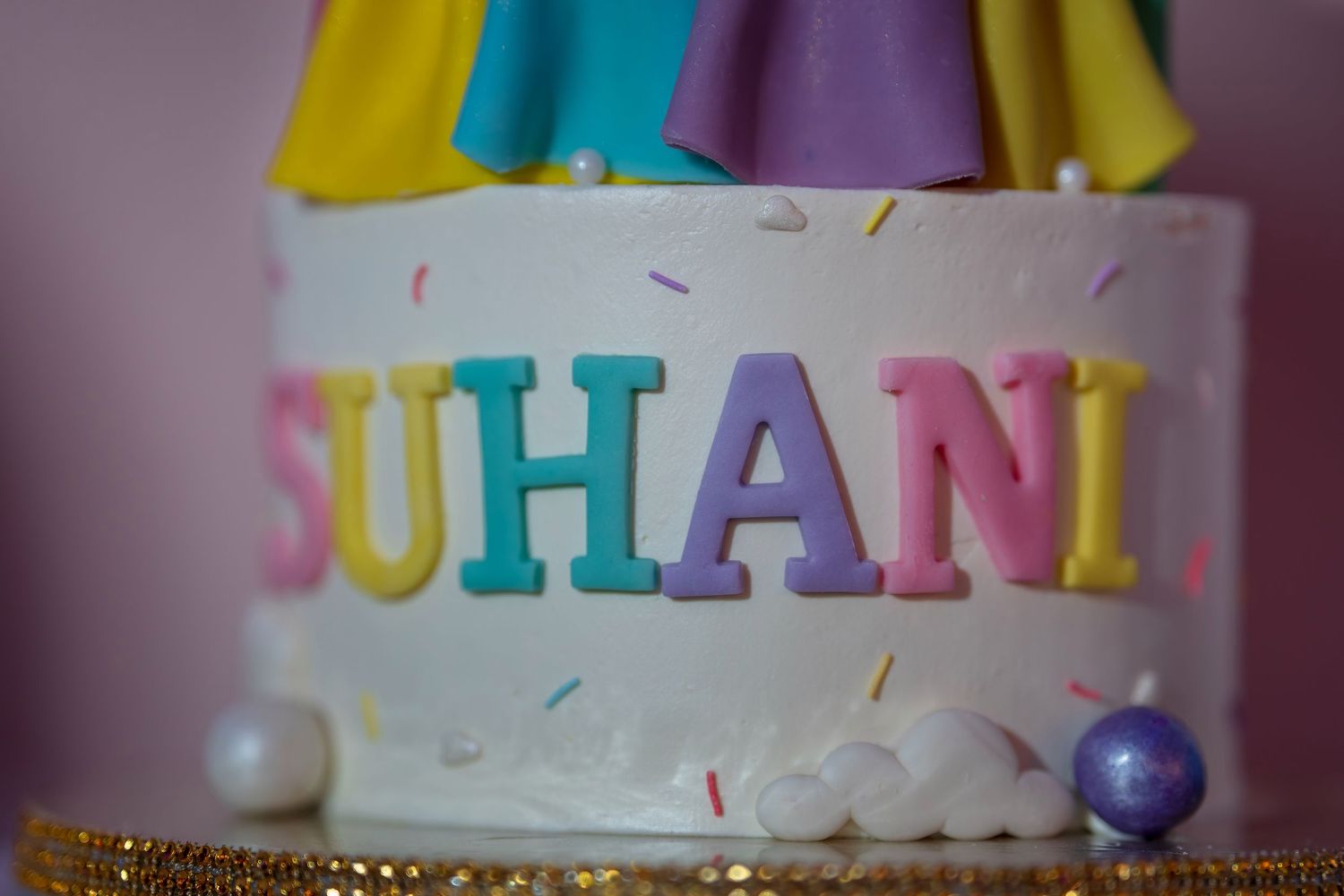 A white birthday cake decorated with colorful letters spelling 'SUHANI' and rainbow sprinkles.