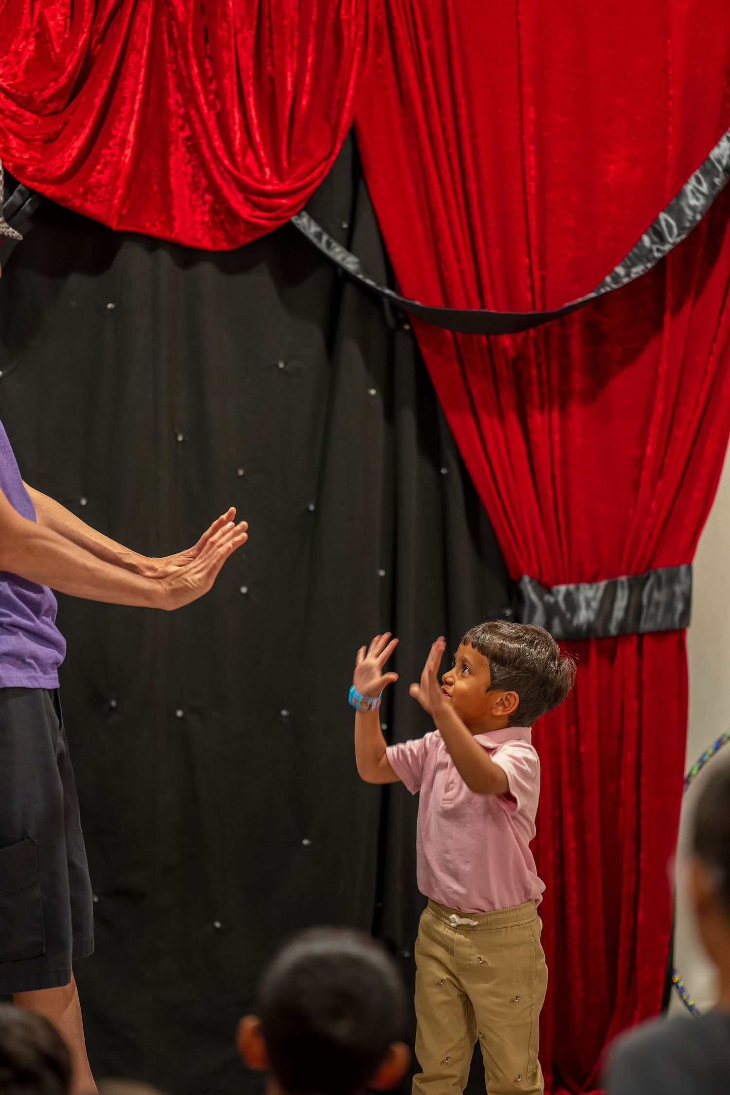 A child reaches upward during a performance against a backdrop of red velvet theater curtains.