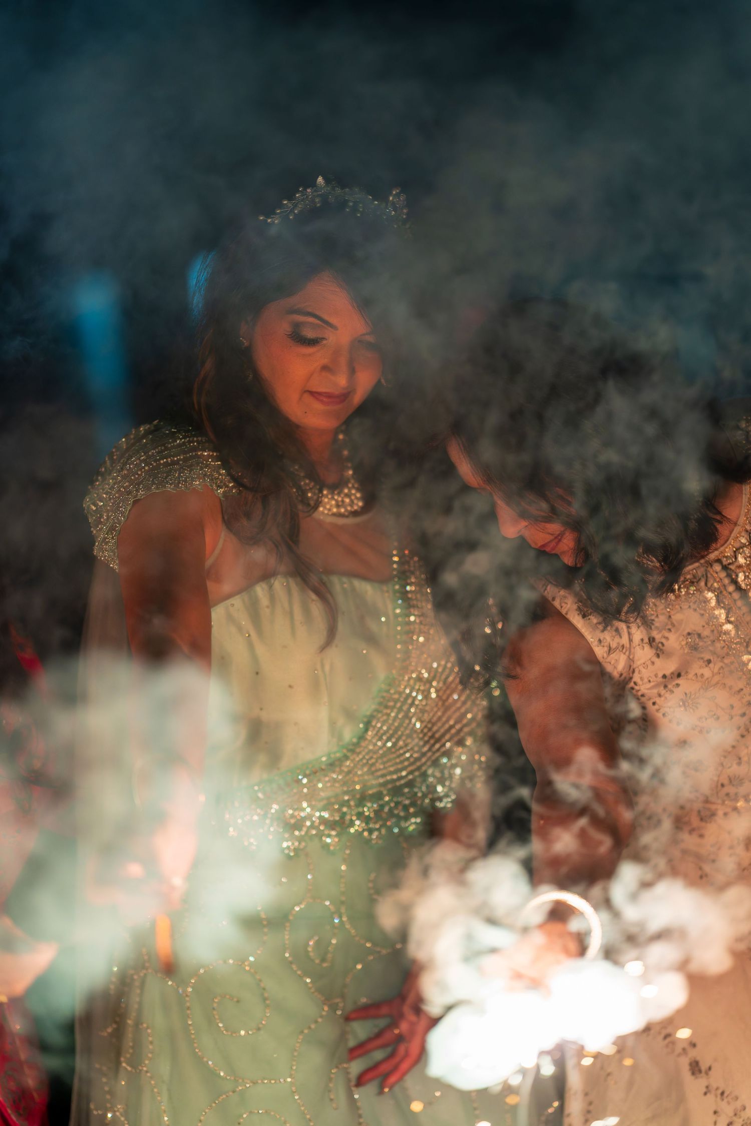 A person in an elegant beaded gown stands among sparkler smoke during an evening event.