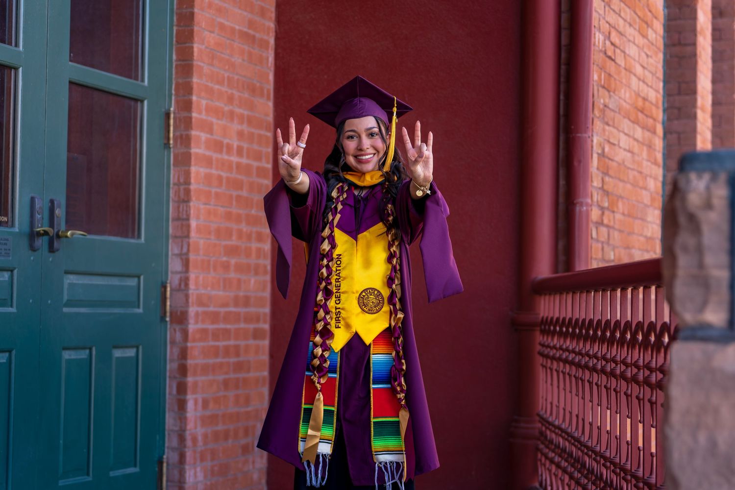 Graduate in traditional purple regalia and bright stole poses confidently against red brick backdrop.