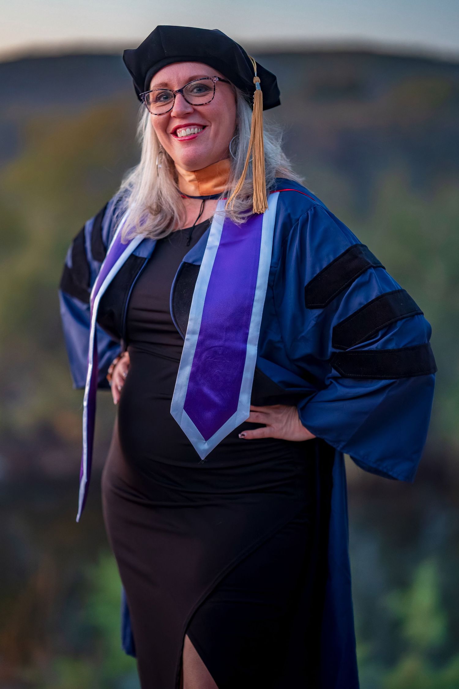 Graduate in traditional academic dress poses outdoors during sunset photo session.