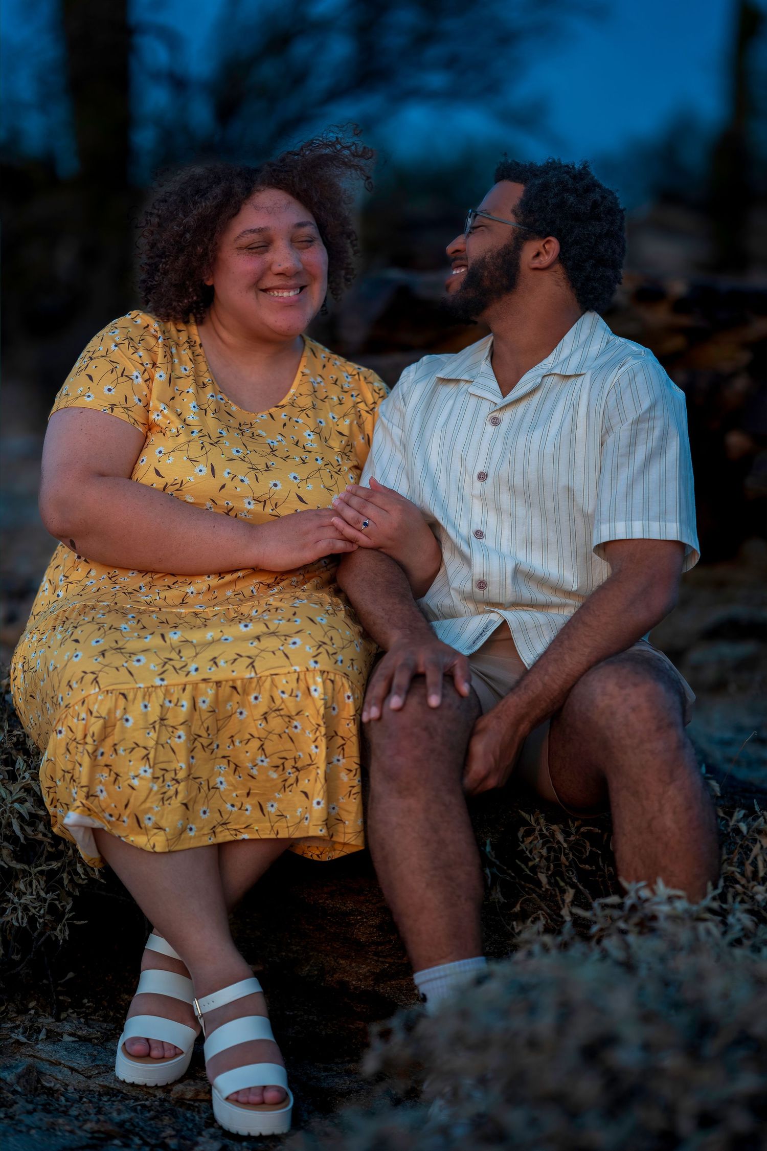Couple sitting together at dusk, woman in yellow dress and man in striped shirt laughing and holding hands.