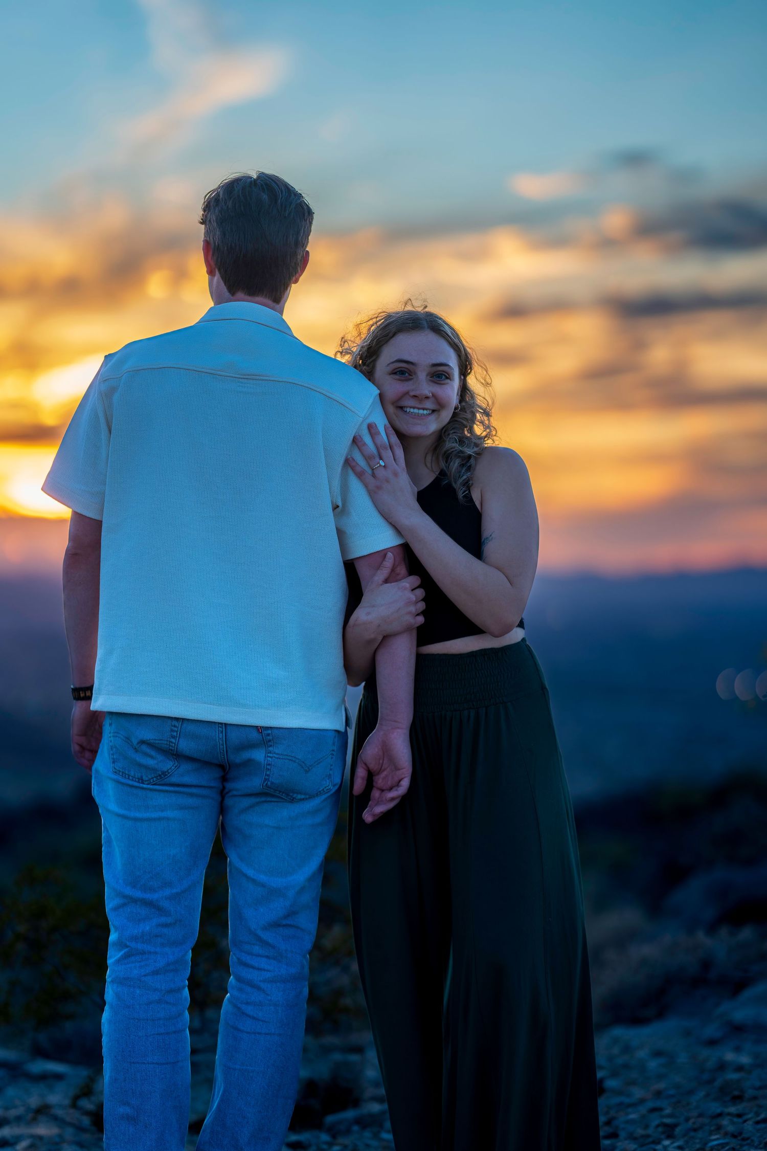 Woman smiling and showing engagement ring while embracing partner facing a vibrant sunset.
