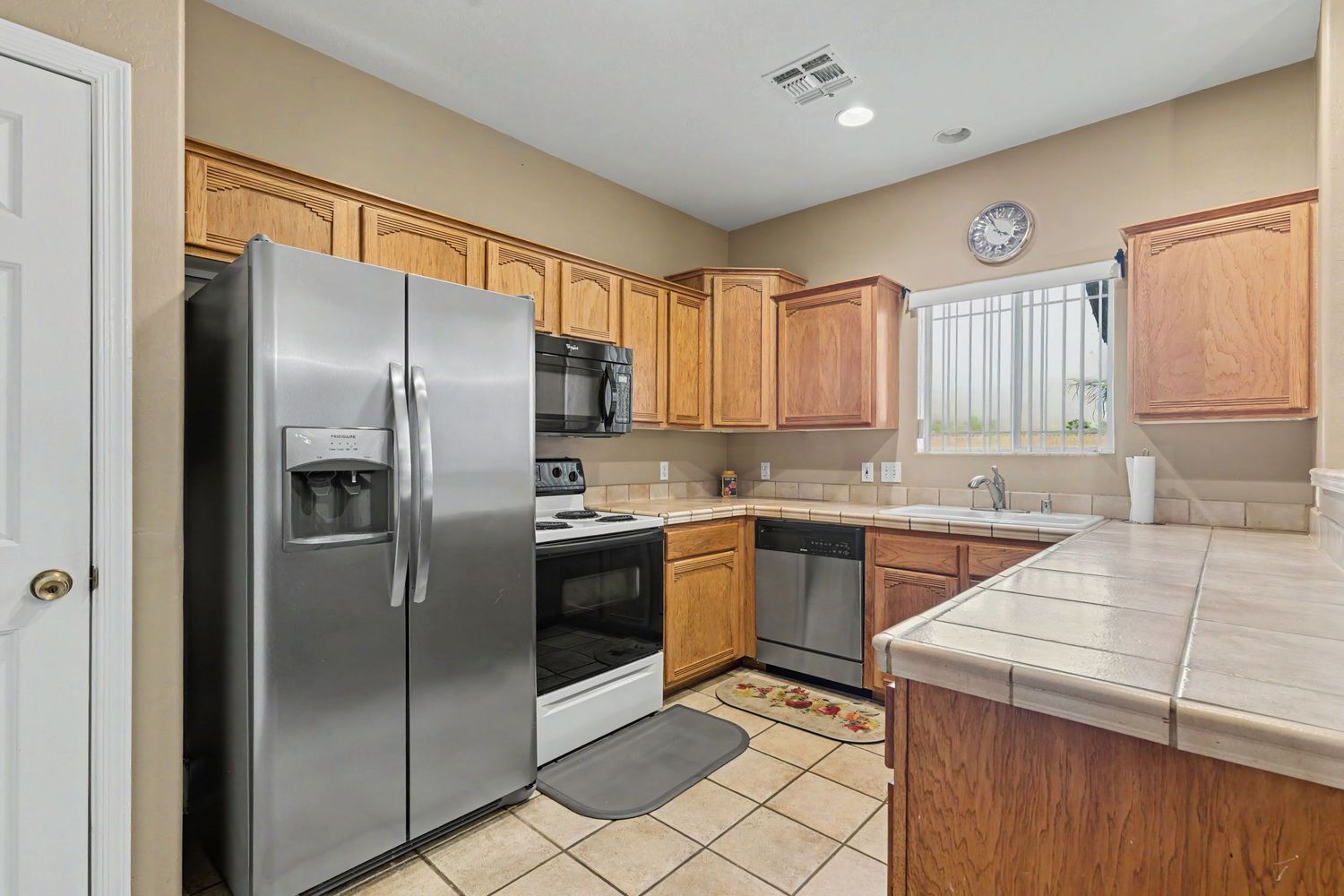 Modern kitchen with stainless steel appliances, oak cabinets, tile countertops, and light tile flooring in a bright home.