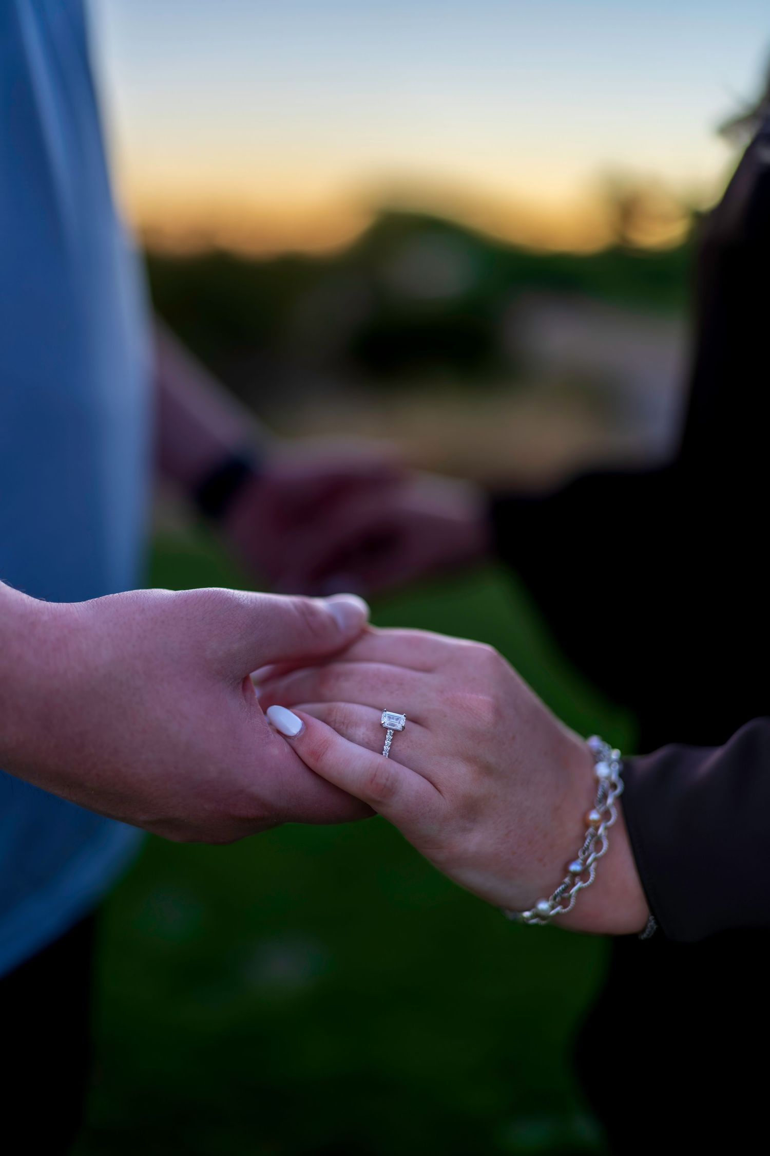 Close-up of a couple holding hands at sunset, showcasing a sparkling diamond engagement ring and silver bracelet.