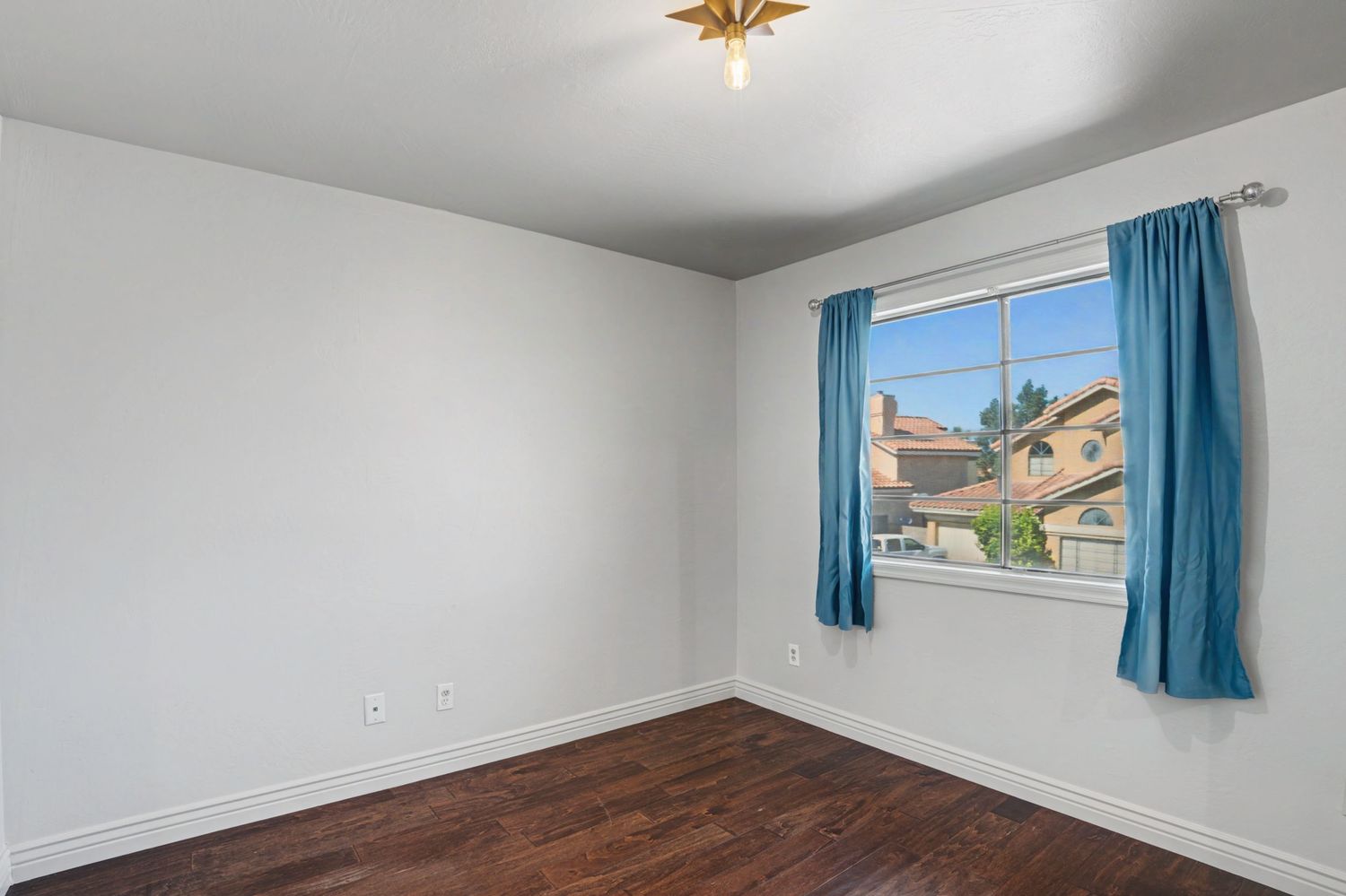 Empty bedroom with dark hardwood floors, white walls, and a window with blue curtains overlooking neighborhood.