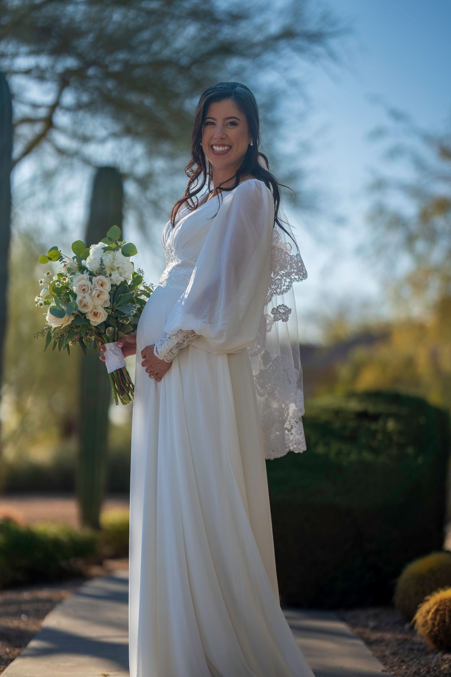 Smiling bride in white long-sleeve lace gown holding elegant floral bouquet outdoors in desert garden setting.