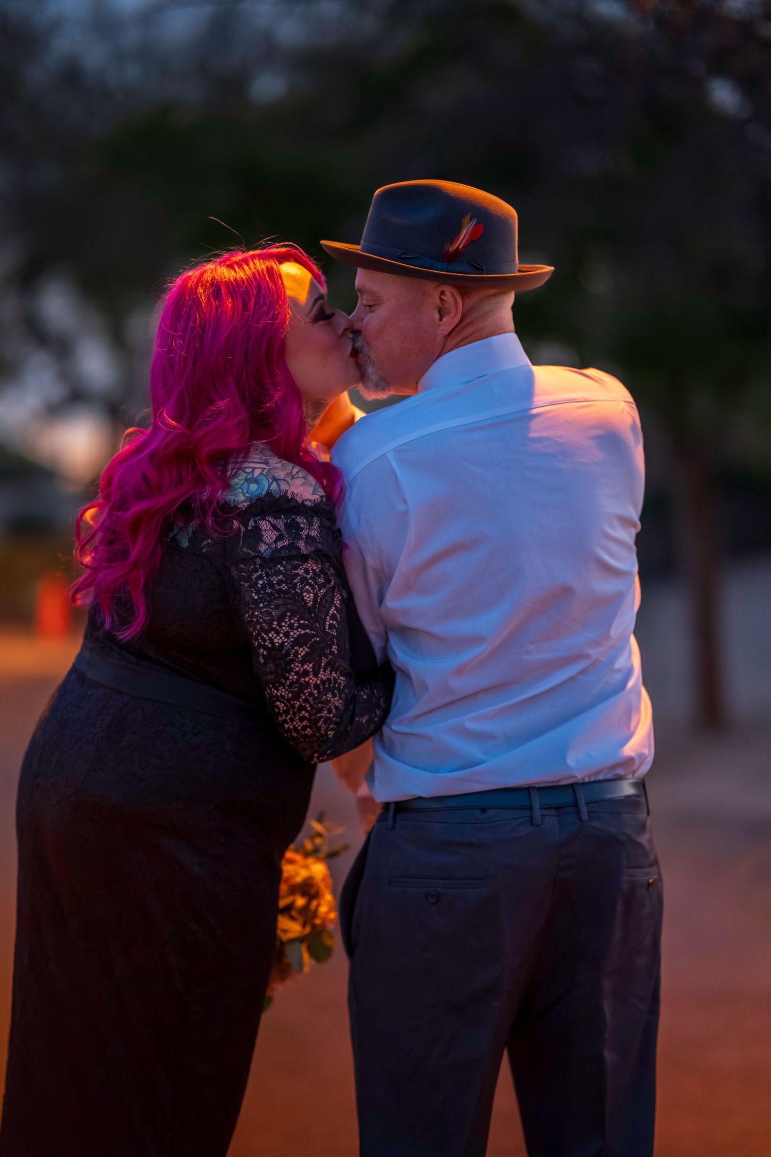Couple sharing a romantic kiss outdoors at dusk, woman with vibrant pink hair in black lace dress, man wearing fedora.