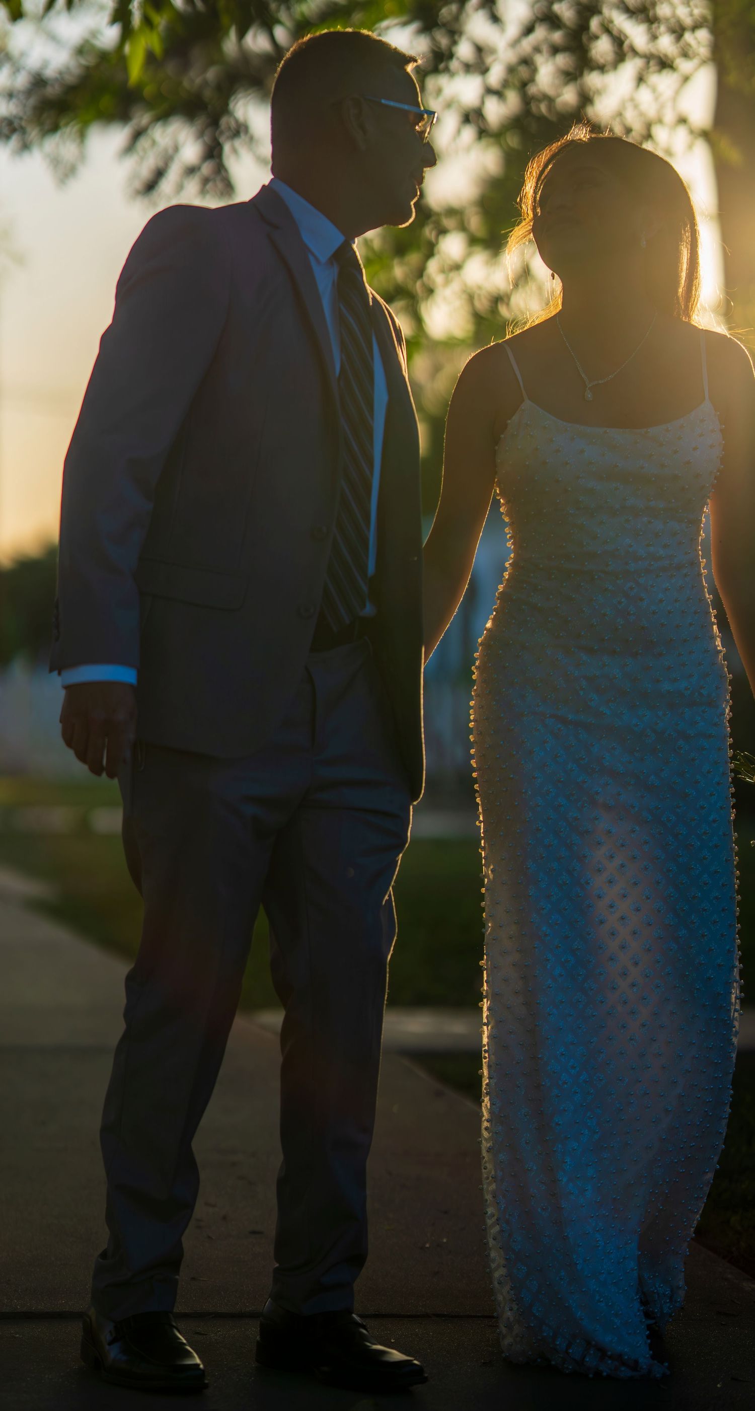 Elegant couple in formal attire holding hands while walking in golden sunset light along a tree-lined path.