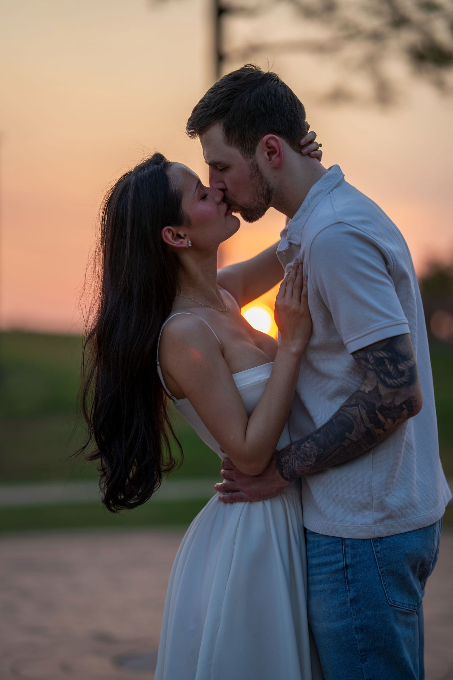 Couple sharing a passionate kiss at sunset, man has tattooed arm, woman wears white dress.