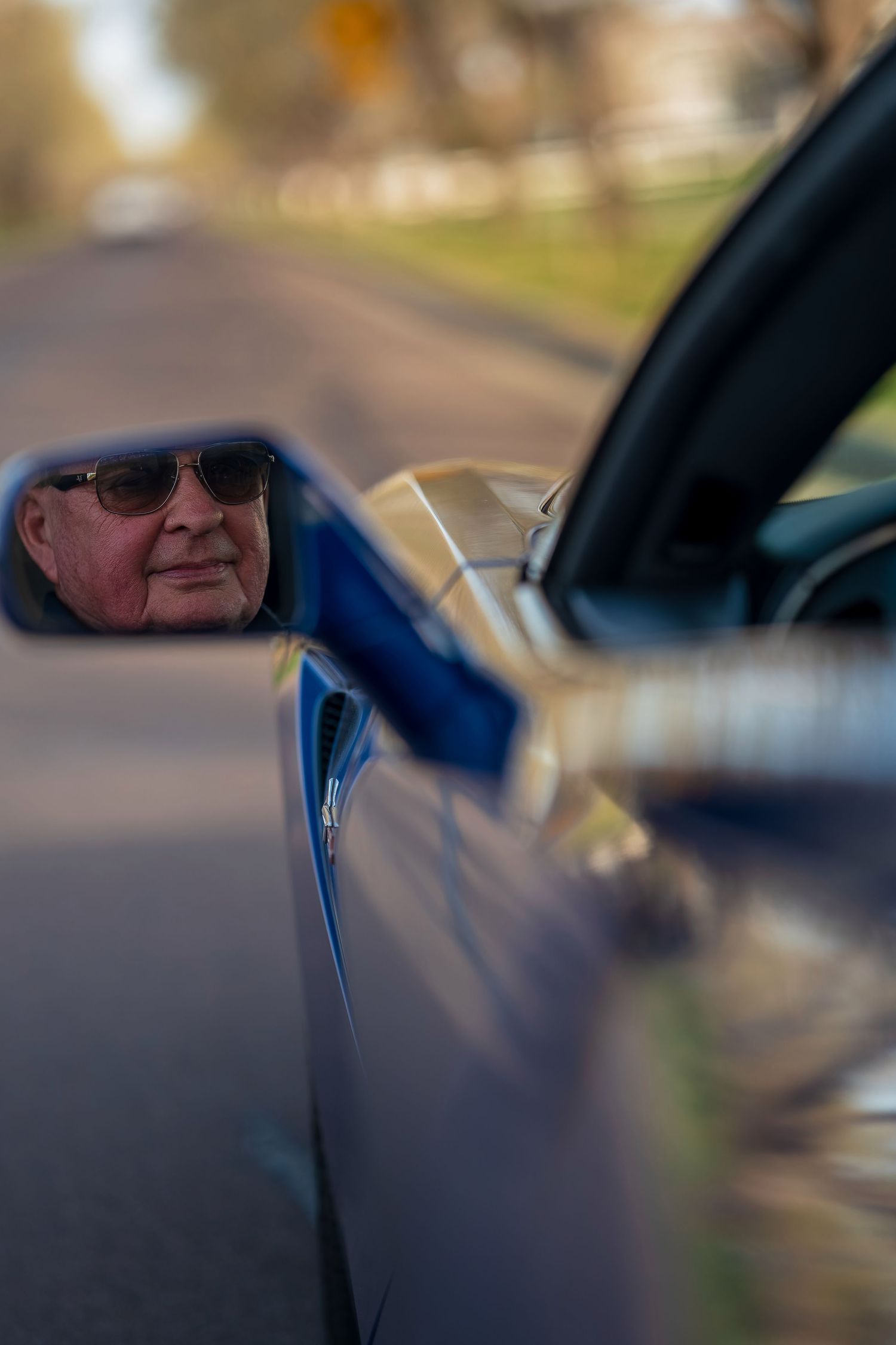 Elderly man wearing sunglasses reflected in the side mirror of a blue sports car on a tree-lined road.