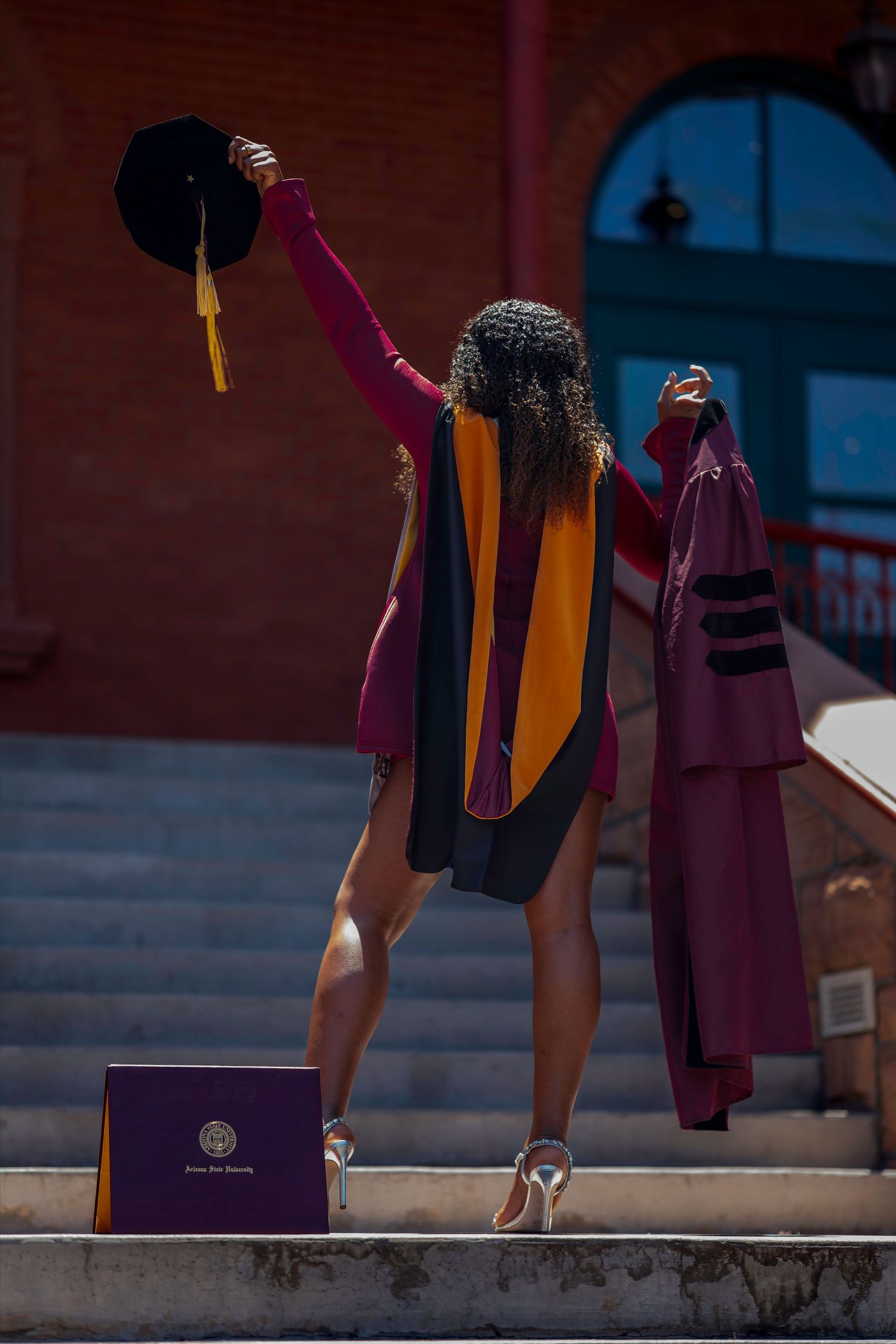 Graduate celebrates on steps holding cap and gown with diploma, wearing maroon and gold stole.
