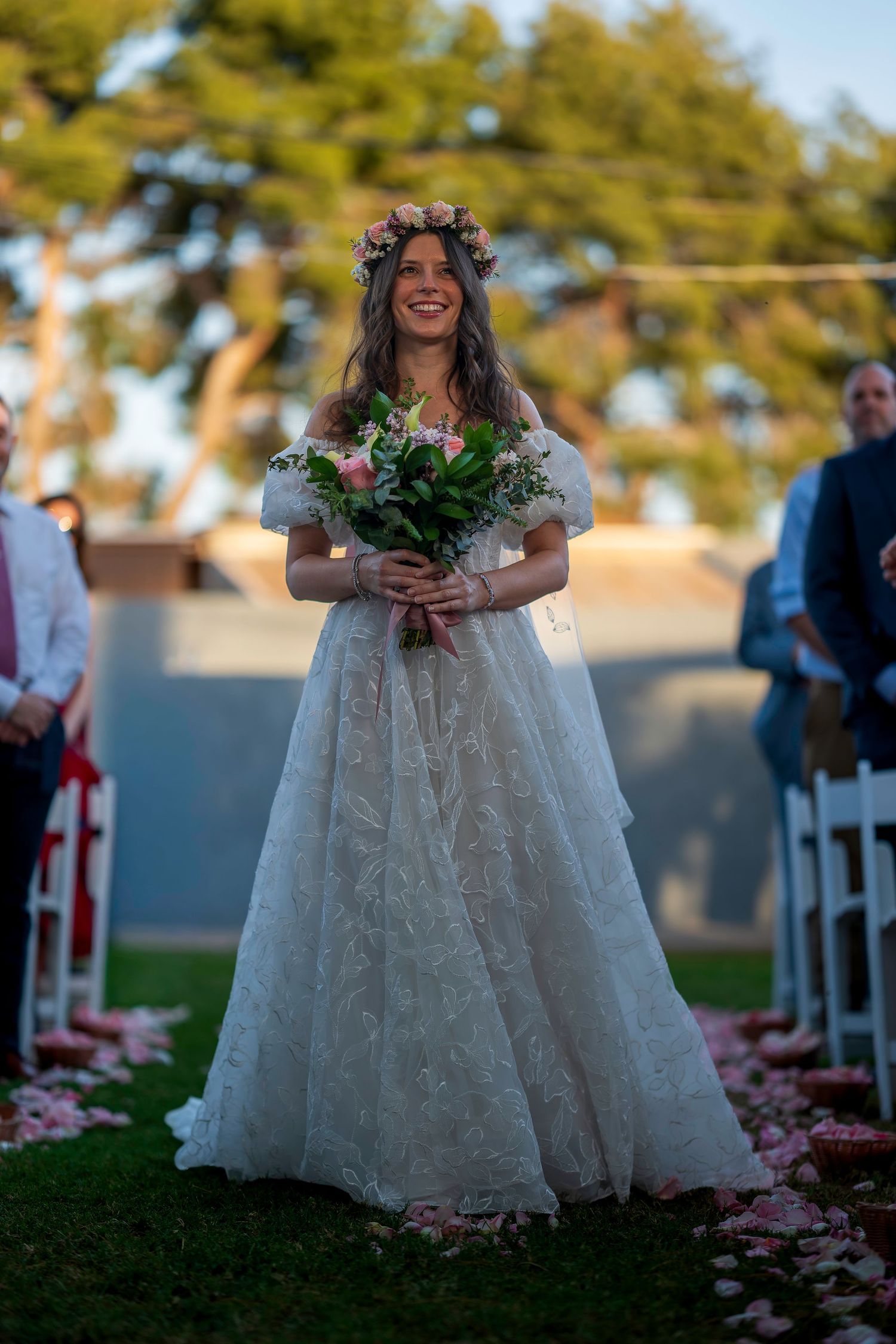 Smiling bride in lace off-shoulder gown with floral crown walks down outdoor aisle holding pink bouquet.