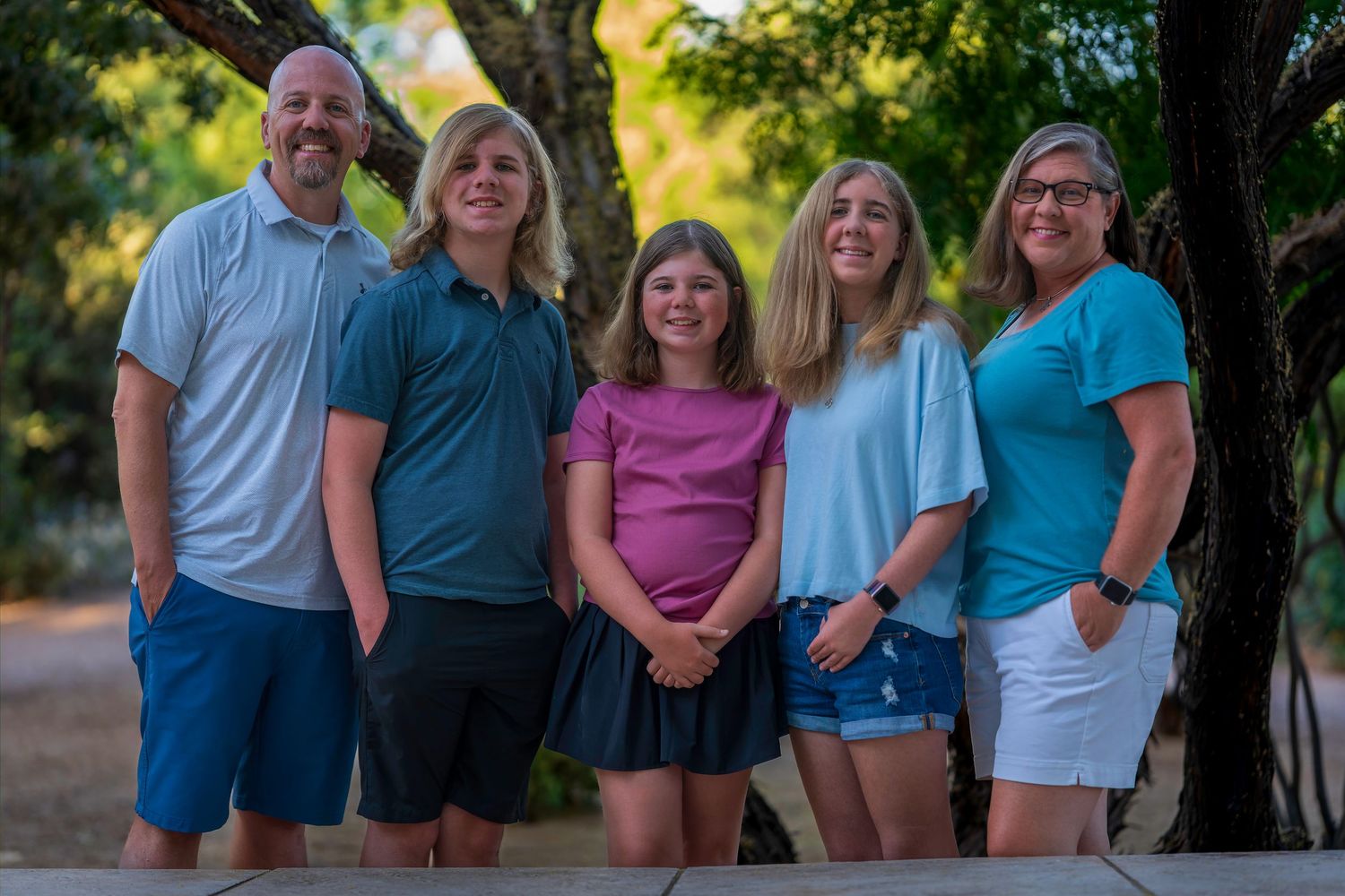 Family of five smiling outdoors under shaded trees, wearing casual summer clothing on a sunny day.