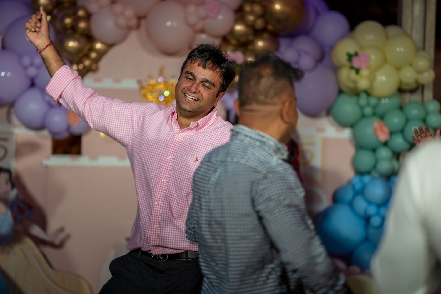 People dancing and celebrating under pastel balloon arrangements at an indoor party.