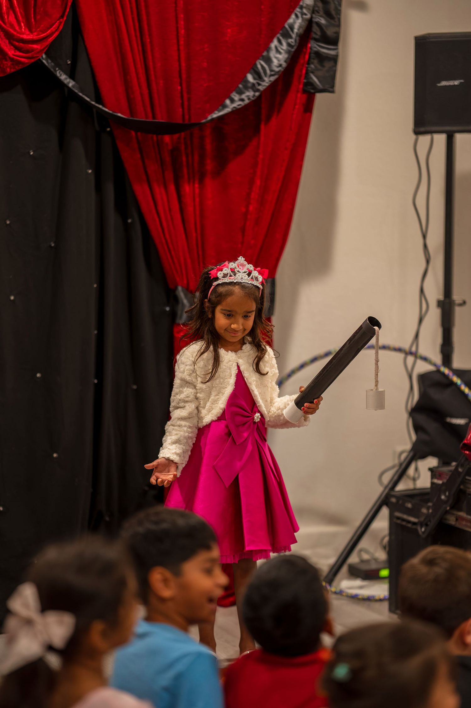 A stage performance with red velvet curtains and audience members visible in the background.