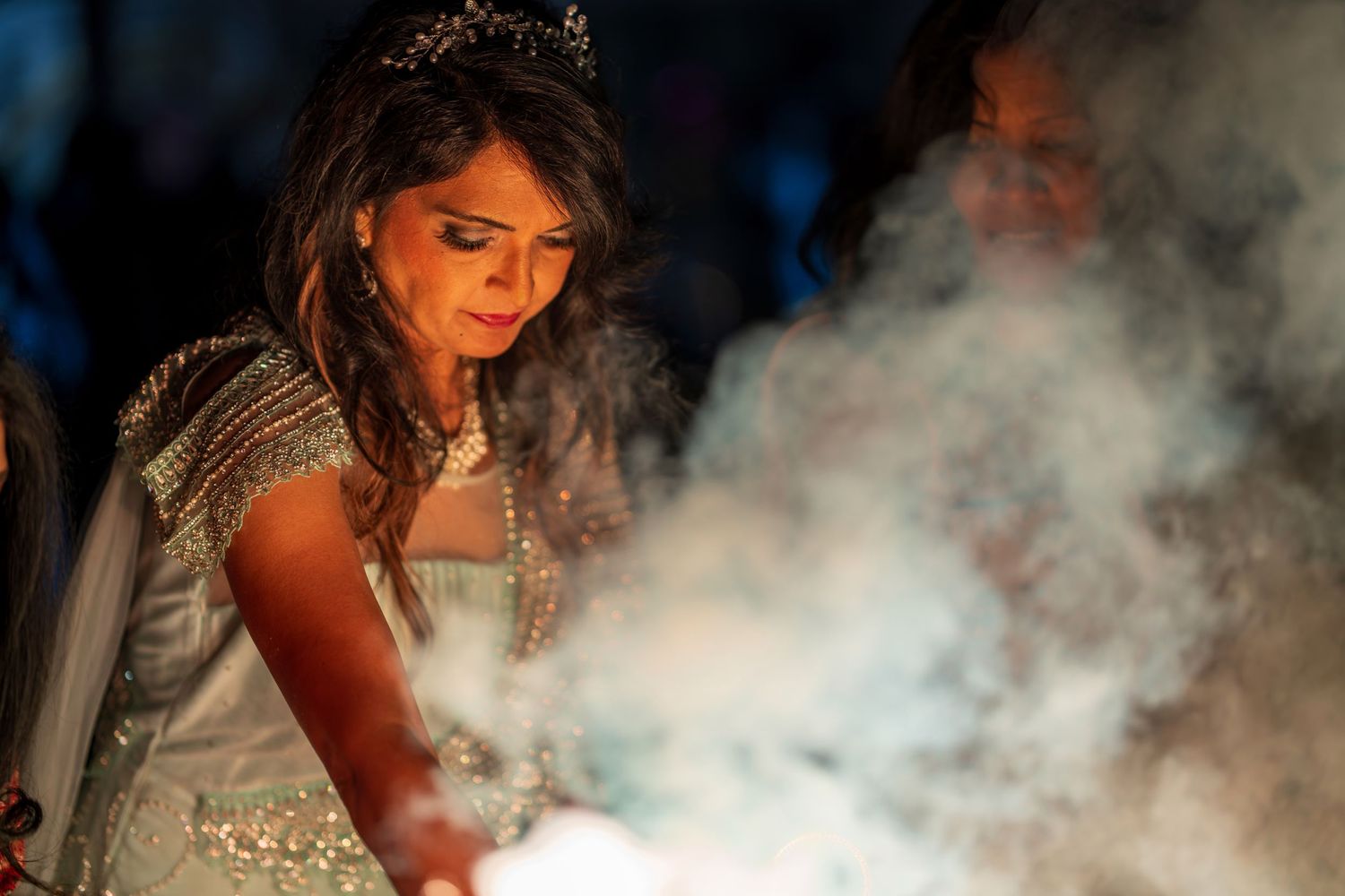A person in a sparkly white dress holds sparklers amidst swirling smoke at a celebration.