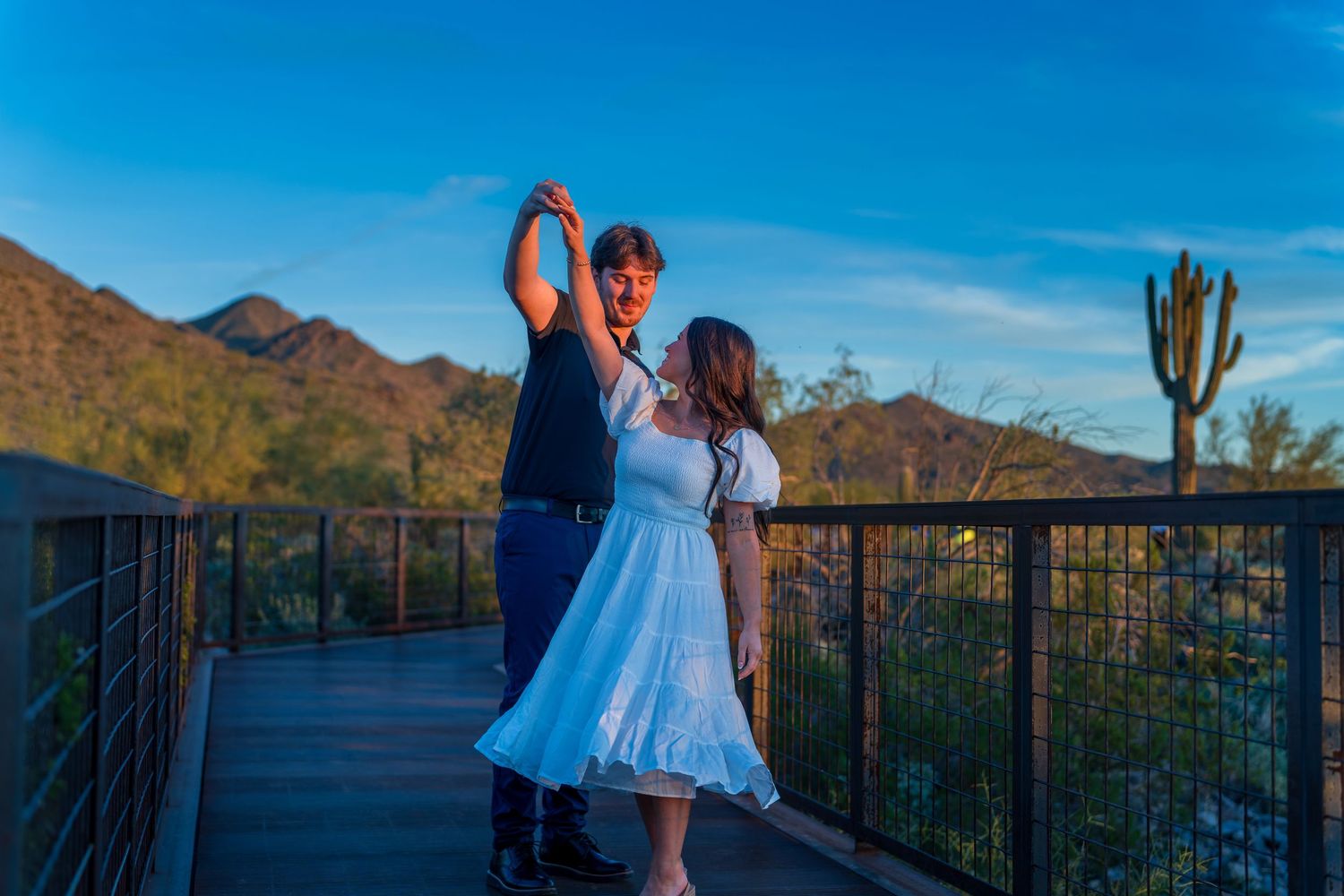 A couple poses together on a deck with desert mountains and saguaro cactus in view.
