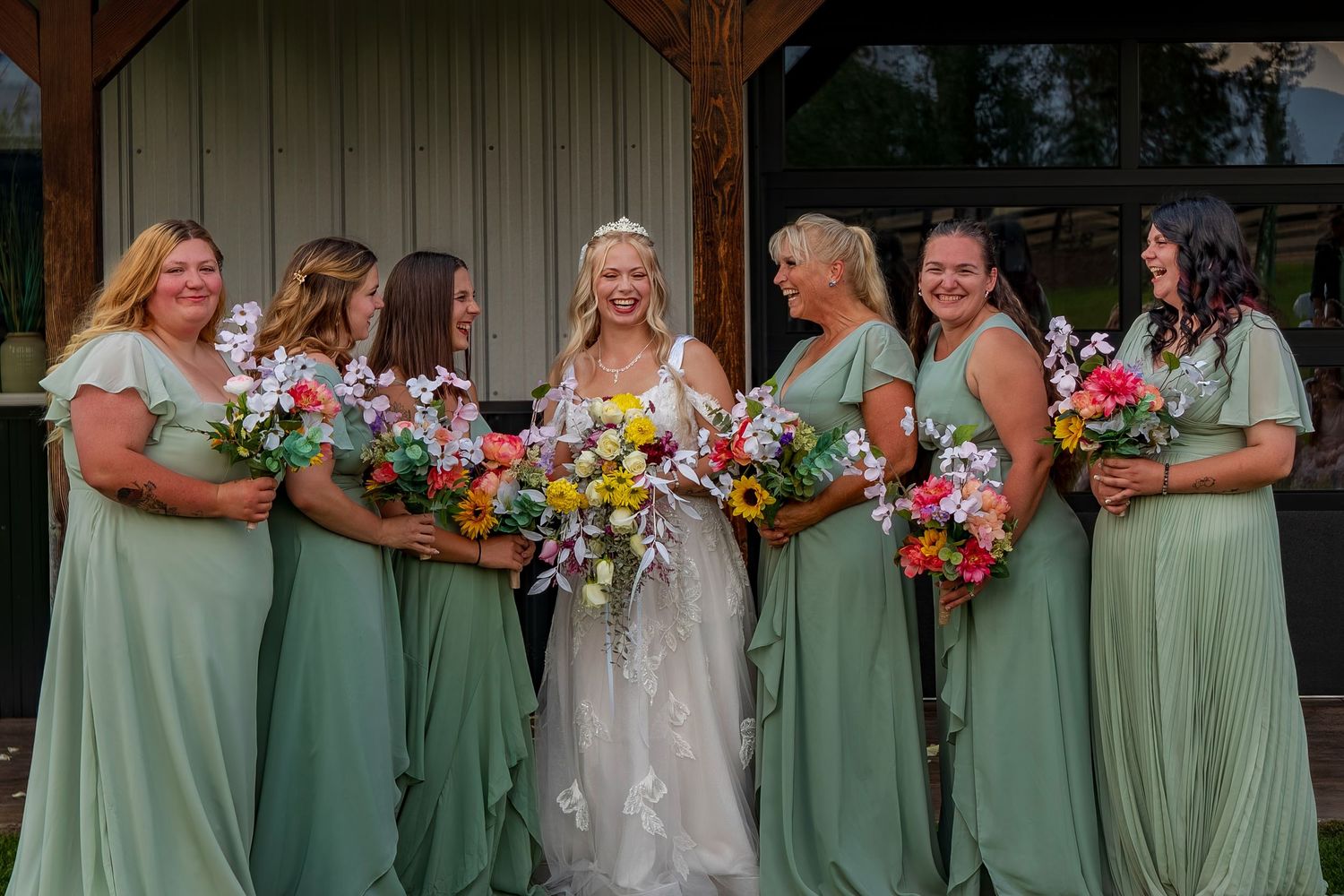 Bridesmaids in sage green dresses pose with colorful bouquets at a rustic venue.