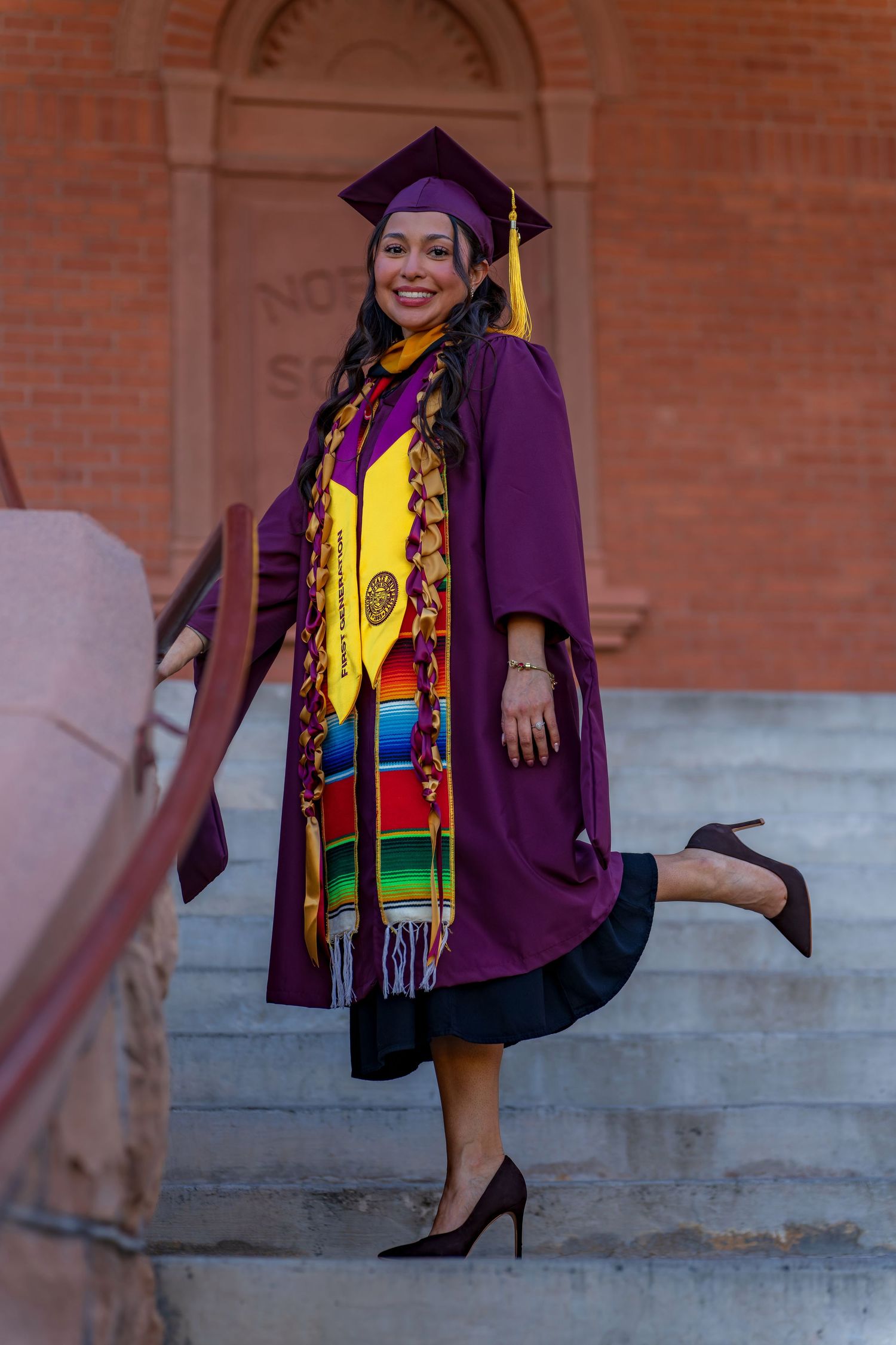 Graduate in purple academic dress and colorful stole poses playfully on concrete steps.