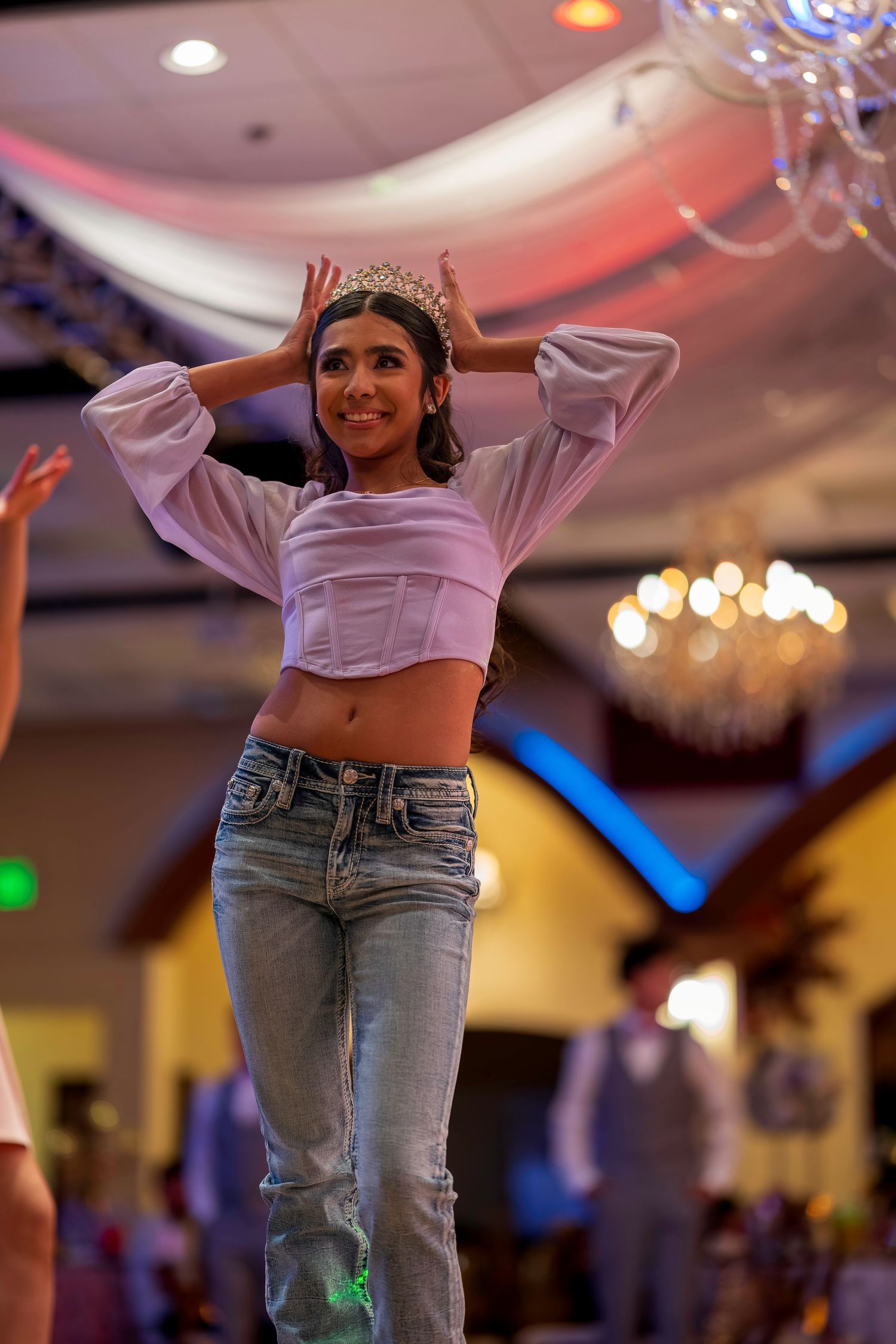Young woman wearing a crown and white crop top with jeans dancing at an elegant event venue.