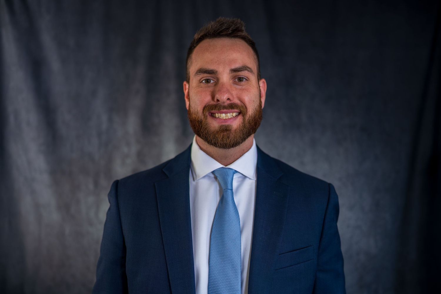 Professional headshot of person in navy suit and light blue tie against dark background.