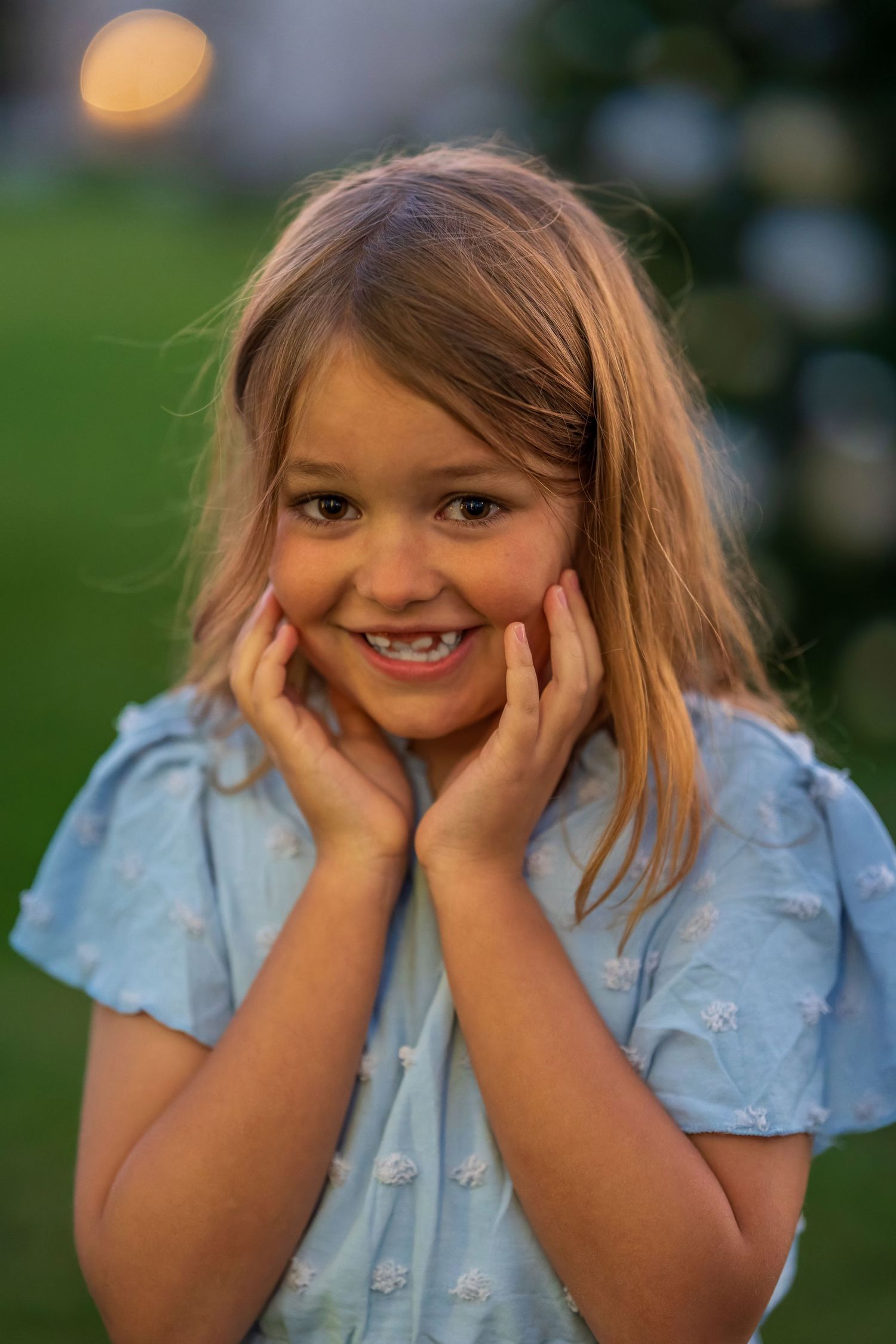 Young child in light blue dress smiling and holding face with both hands outdoors.