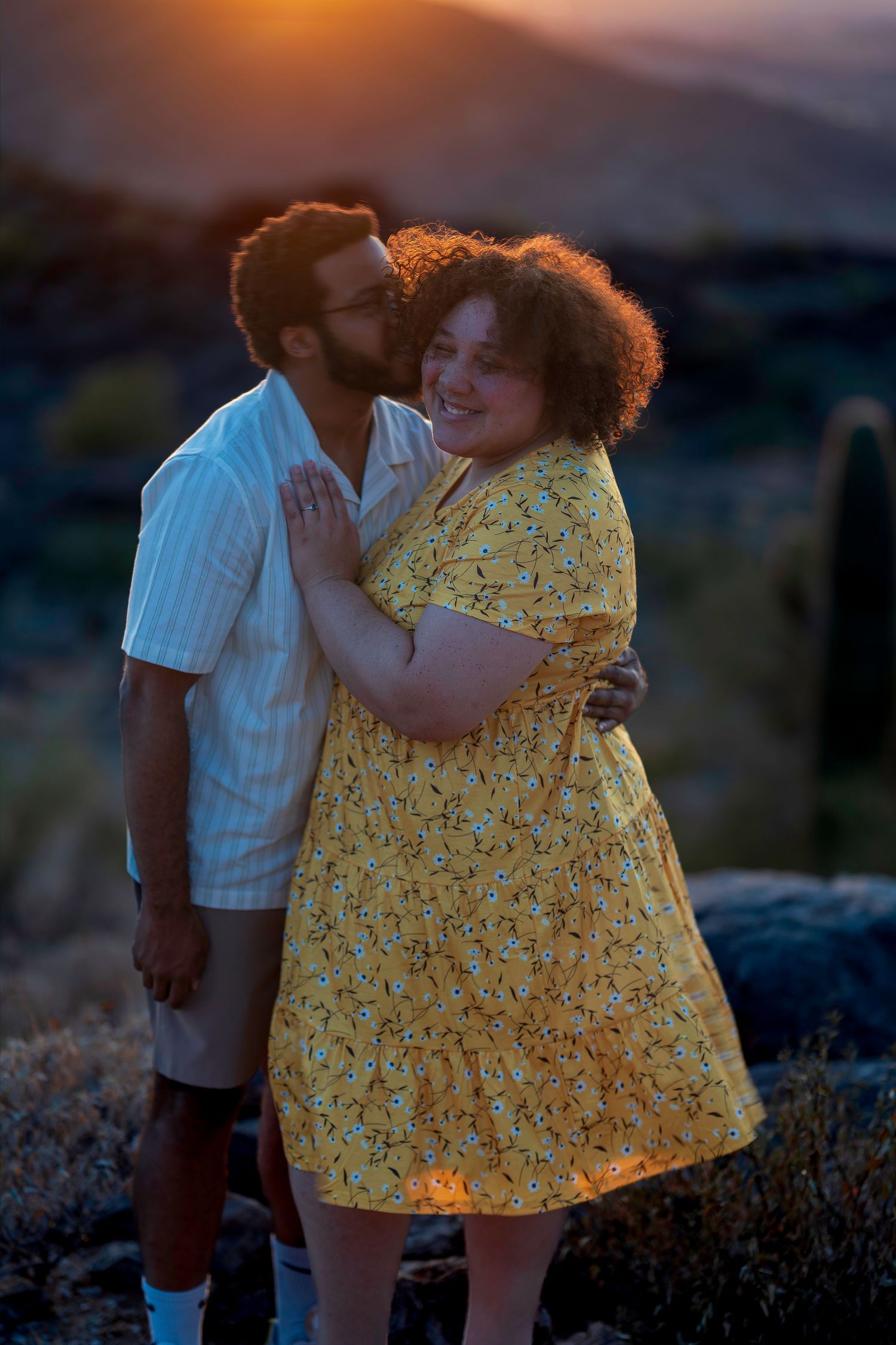 Couple embracing at sunset in desert landscape, woman in yellow floral dress smiling as man kisses her cheek.