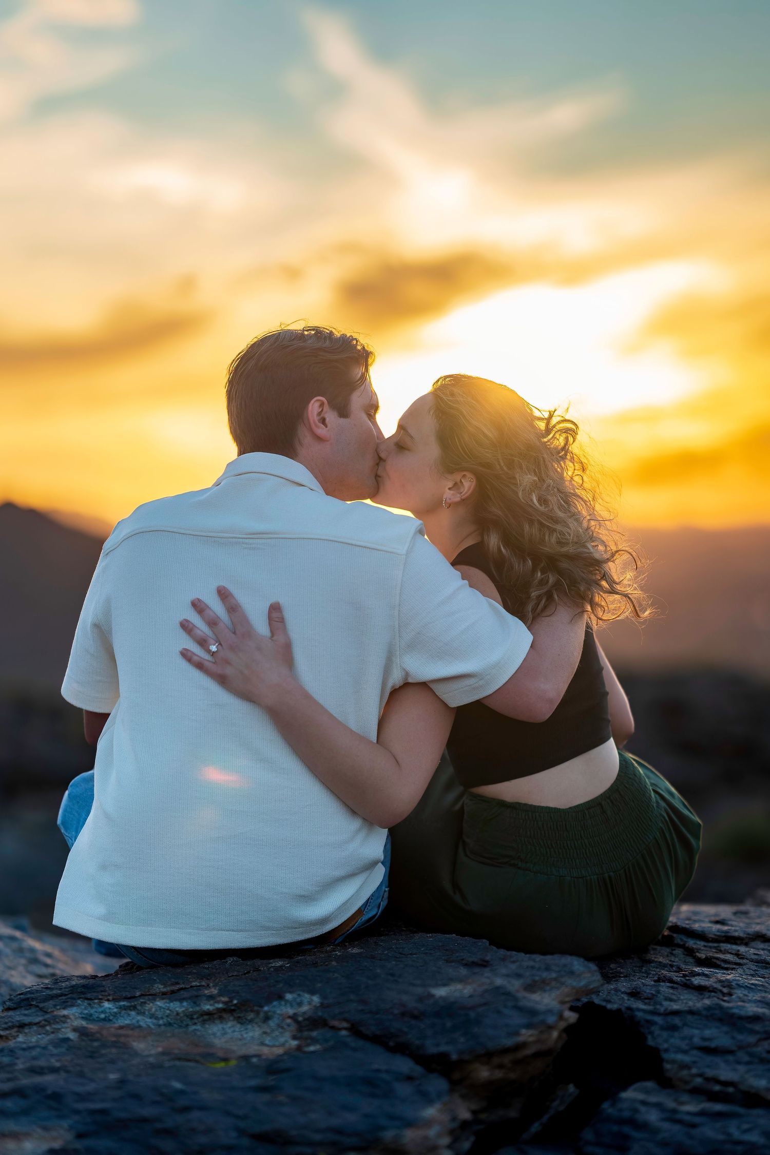 Couple kissing at sunset while sitting on rocks with a vibrant orange and yellow sky behind them.