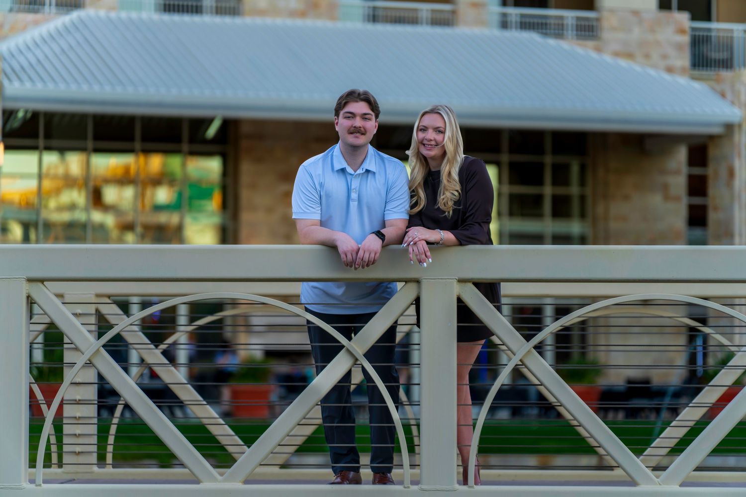 A young couple smiles while leaning on a decorative bridge railing outside a modern stone building.