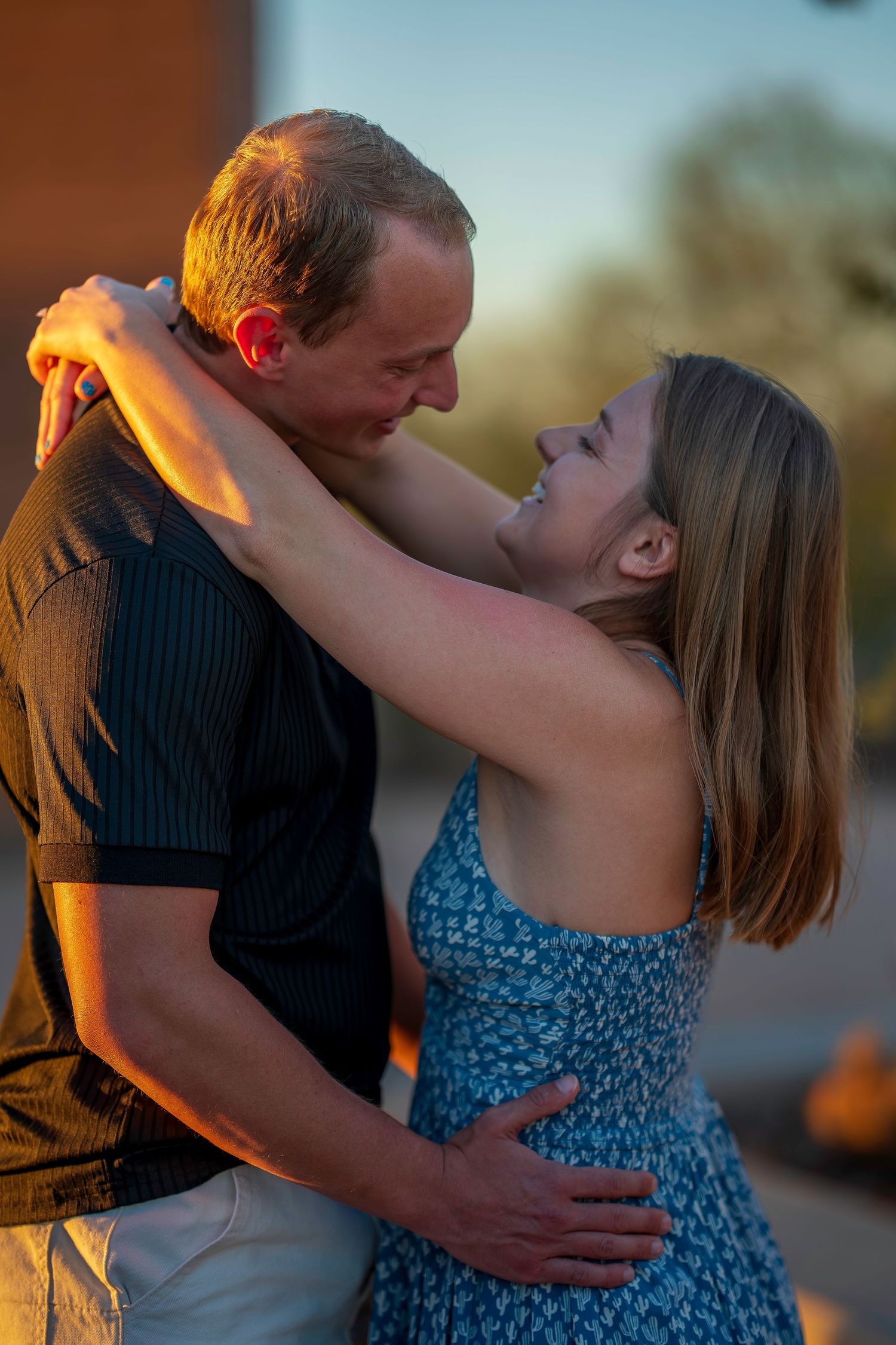 Couple embracing romantically at golden hour sunset, woman in blue floral dress smiling up at man in black shirt.