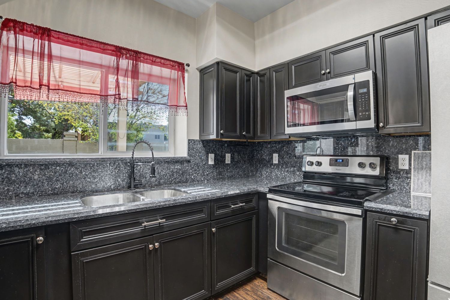 Dark kitchen cabinets with granite countertops, stainless steel appliances, and a window with red curtains.