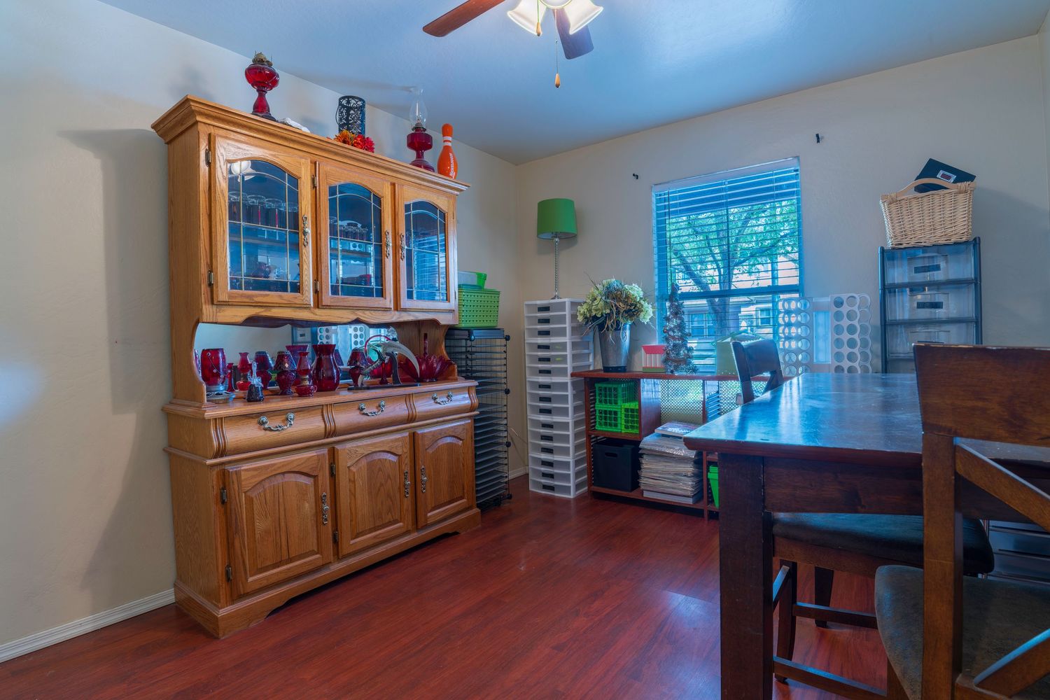 Formal dining room with hardwood floors, oak china hutch displaying red glassware, and ceiling fan.