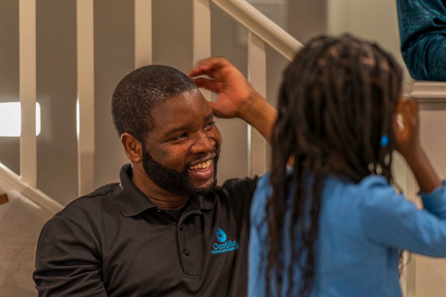 A smiling Black man in a black polo shirt laughs with a child on indoor stairs in a warm home setting.
