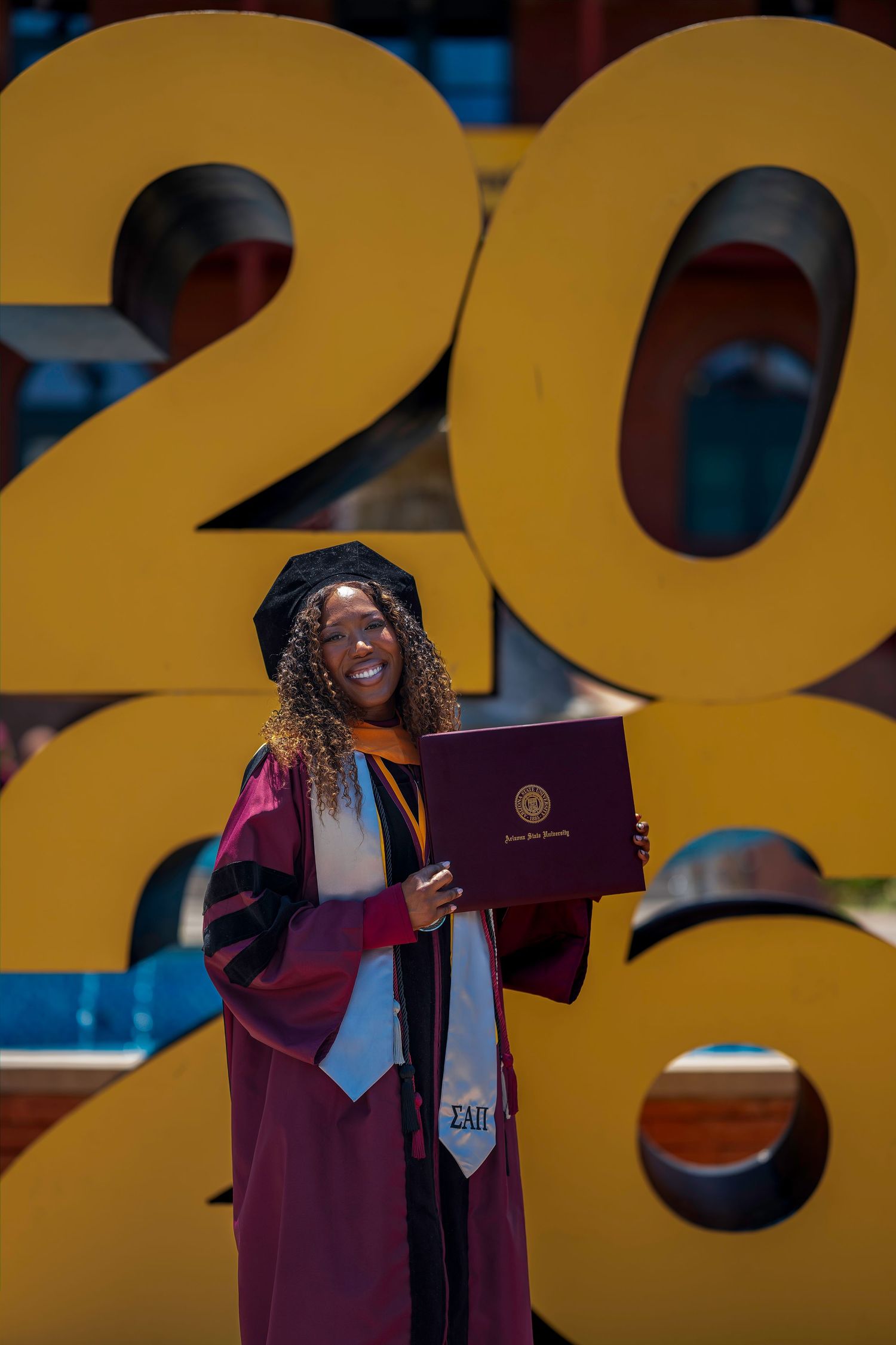 Smiling graduate in maroon regalia holds diploma in front of large gold 2020 numbers outdoors on a sunny day.