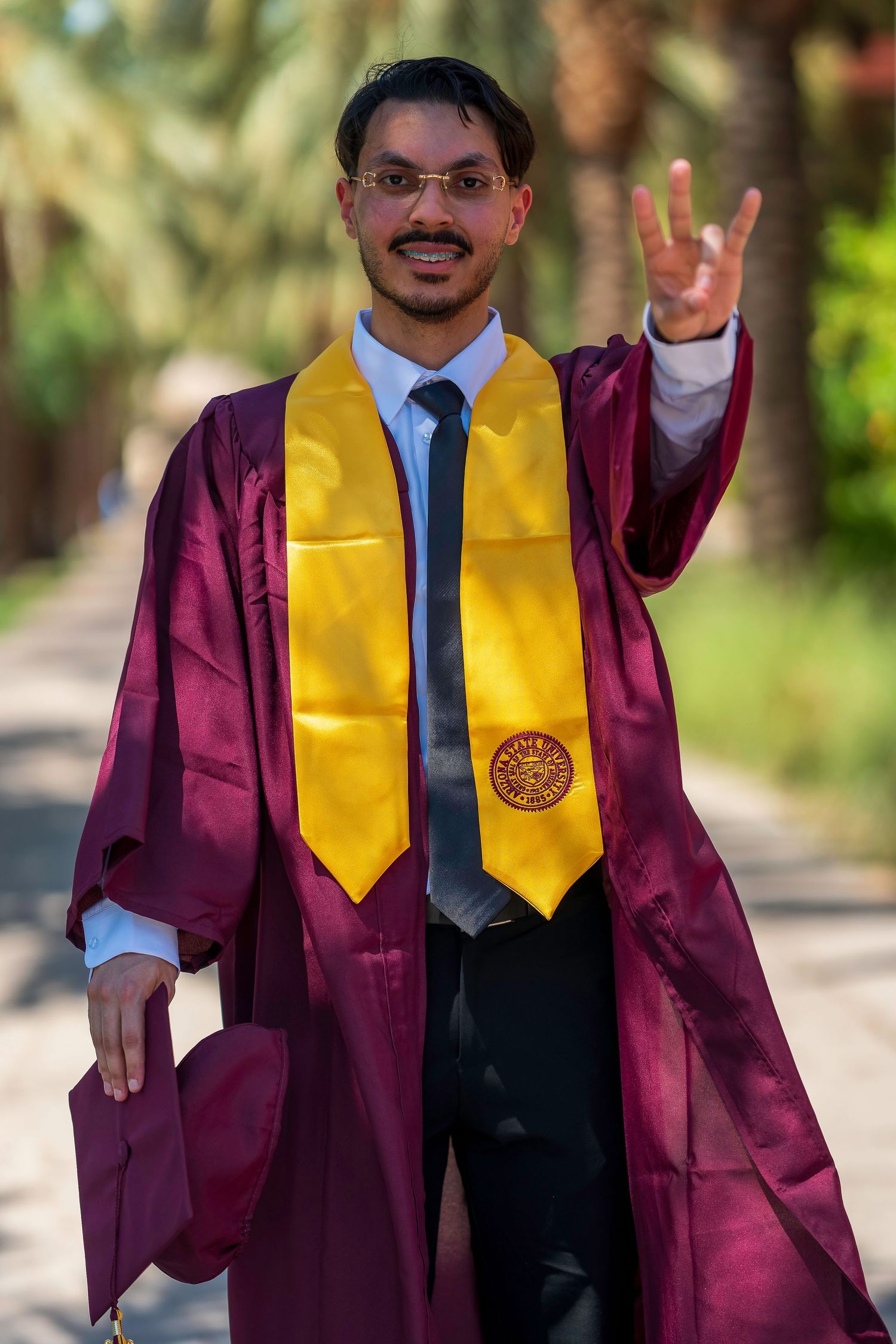 Graduate in maroon robe and gold stole flashing hand sign outdoors.