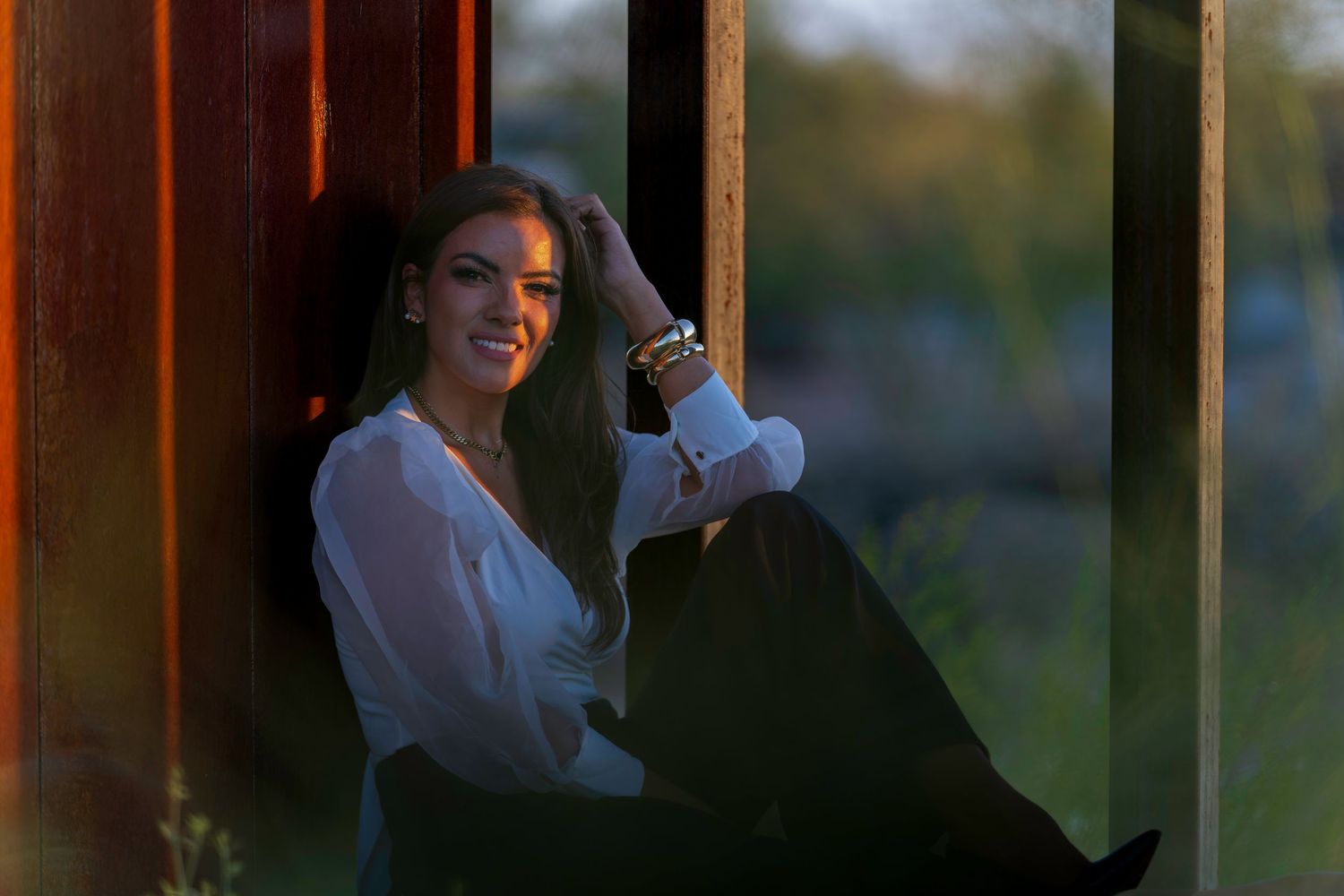 Young woman in white blouse smiling while sitting against a rustic wall in warm golden hour light.