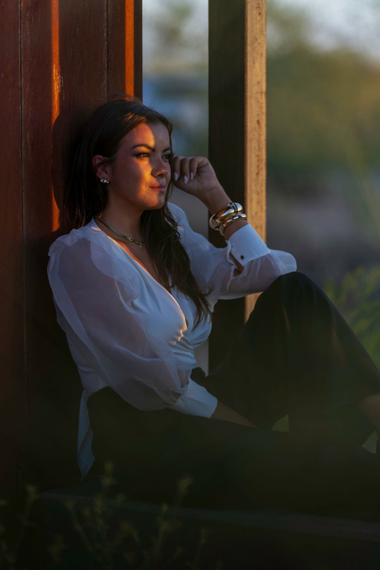 Young woman in white blouse sitting against rustic wall in golden hour light, wearing gold jewelry and dark pants.