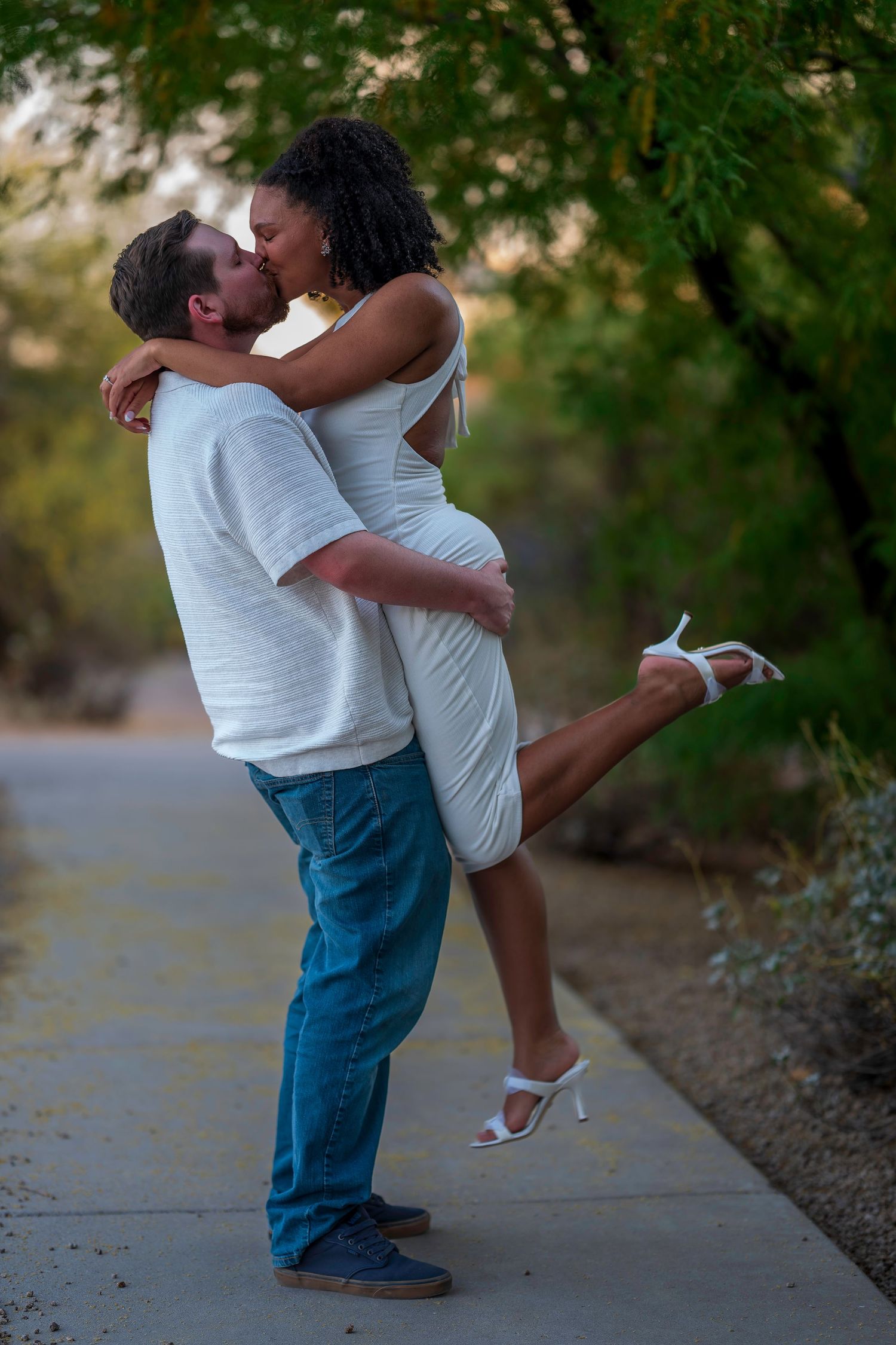 Couple sharing a romantic kiss outdoors, man lifting woman in white dress and heels on a tree-lined path.