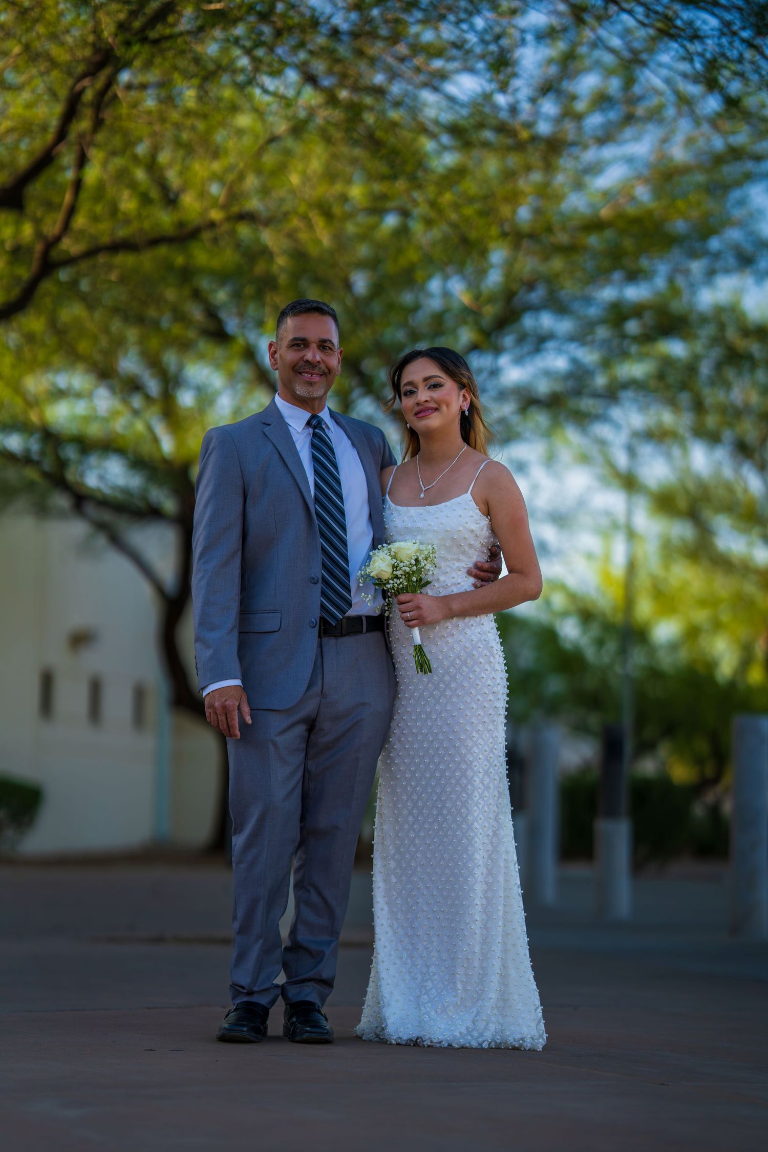 Bride in white beaded gown holding bouquet stands with groom in gray suit under green trees.