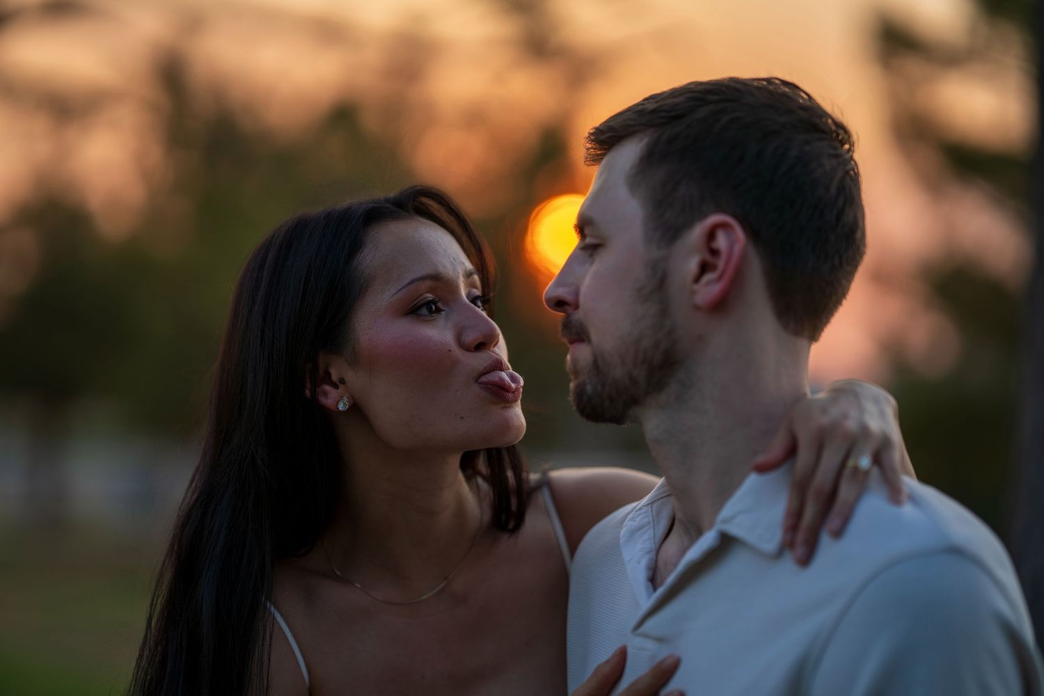 Couple sharing a romantic moment at sunset, woman with dark hair leaning toward bearded man outdoors.