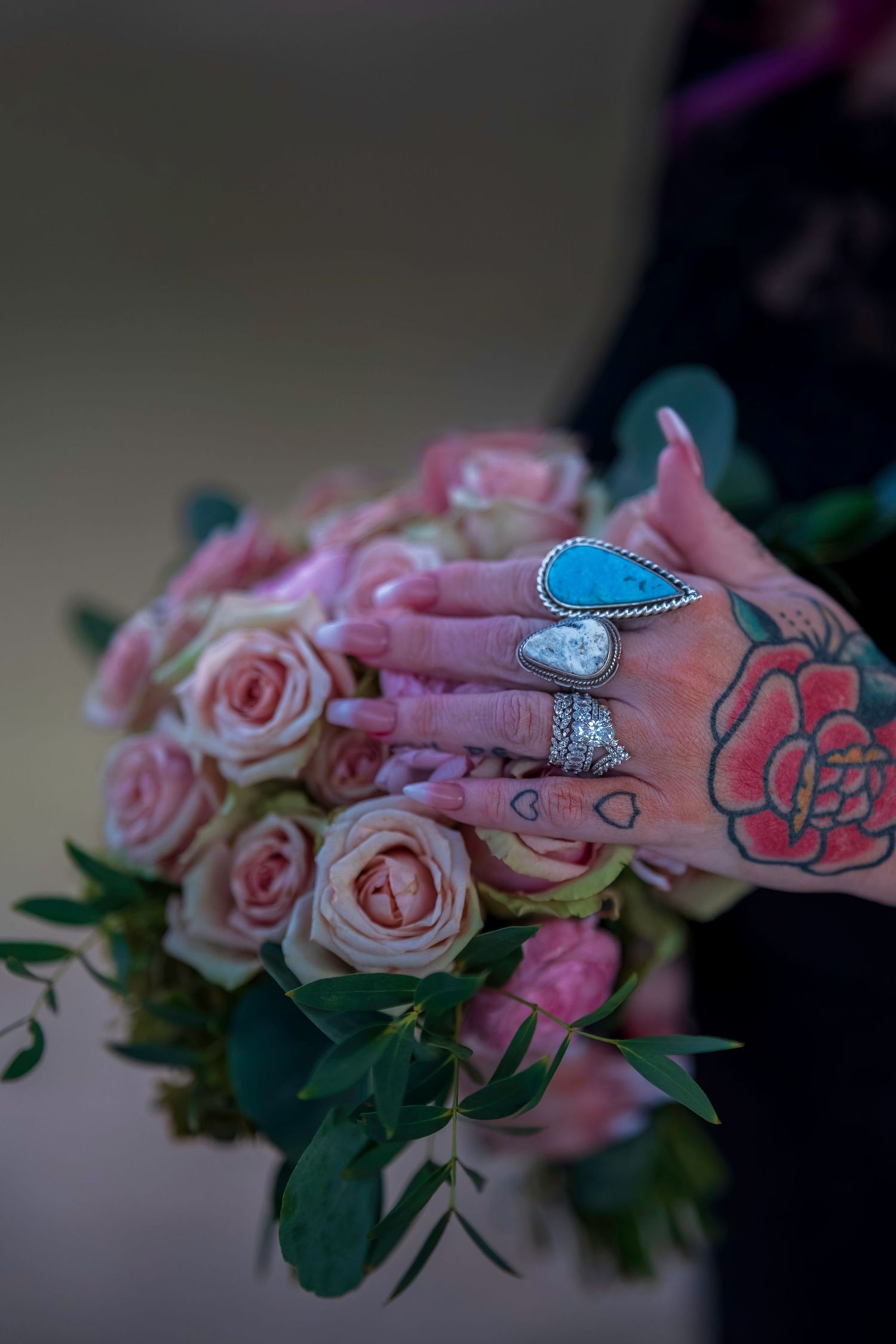 Tattooed hand with turquoise and silver rings resting on a pink rose bouquet with eucalyptus greenery.