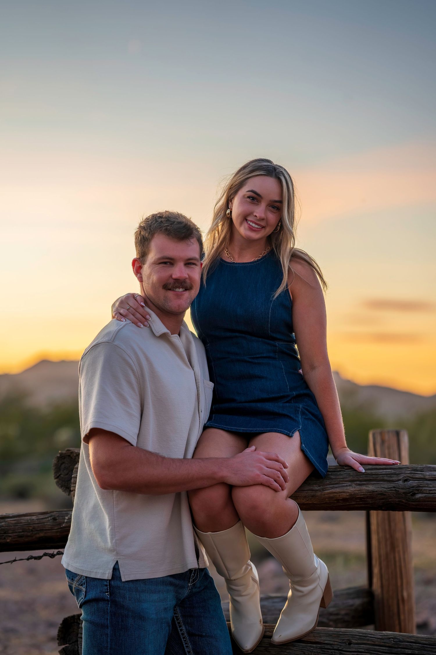 Couple posing at sunset in desert landscape, woman in blue dress and white boots sitting on wooden fence, man in beige shirt.