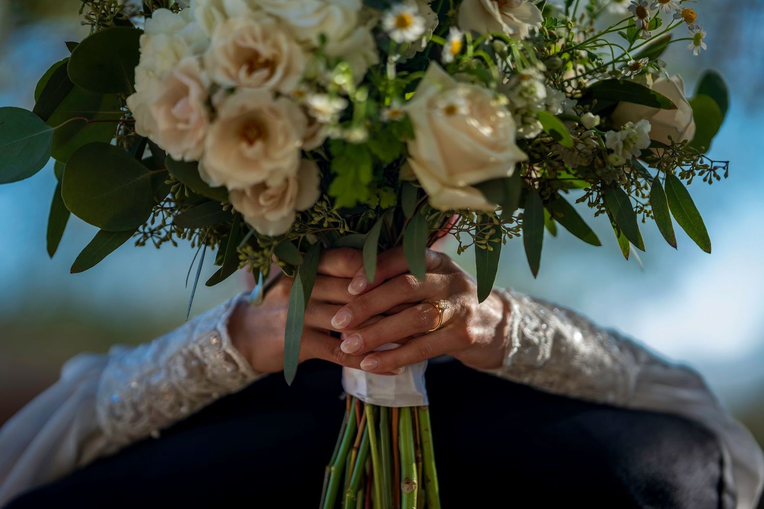 Close-up of bride holding elegant white rose and greenery wedding bouquet with lace sleeve detail.