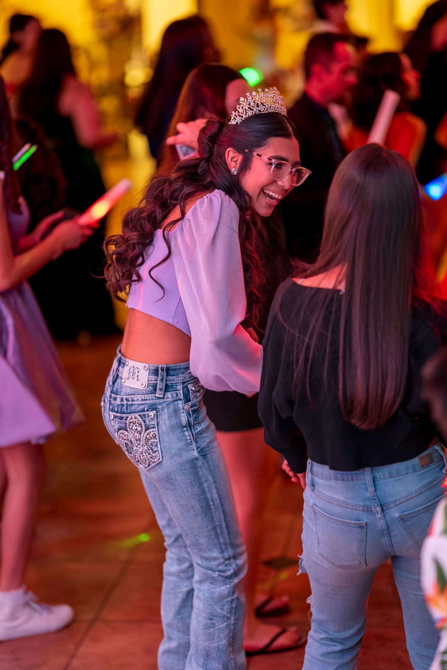 Young woman wearing a tiara and jeans dancing at a colorful party celebration with friends.